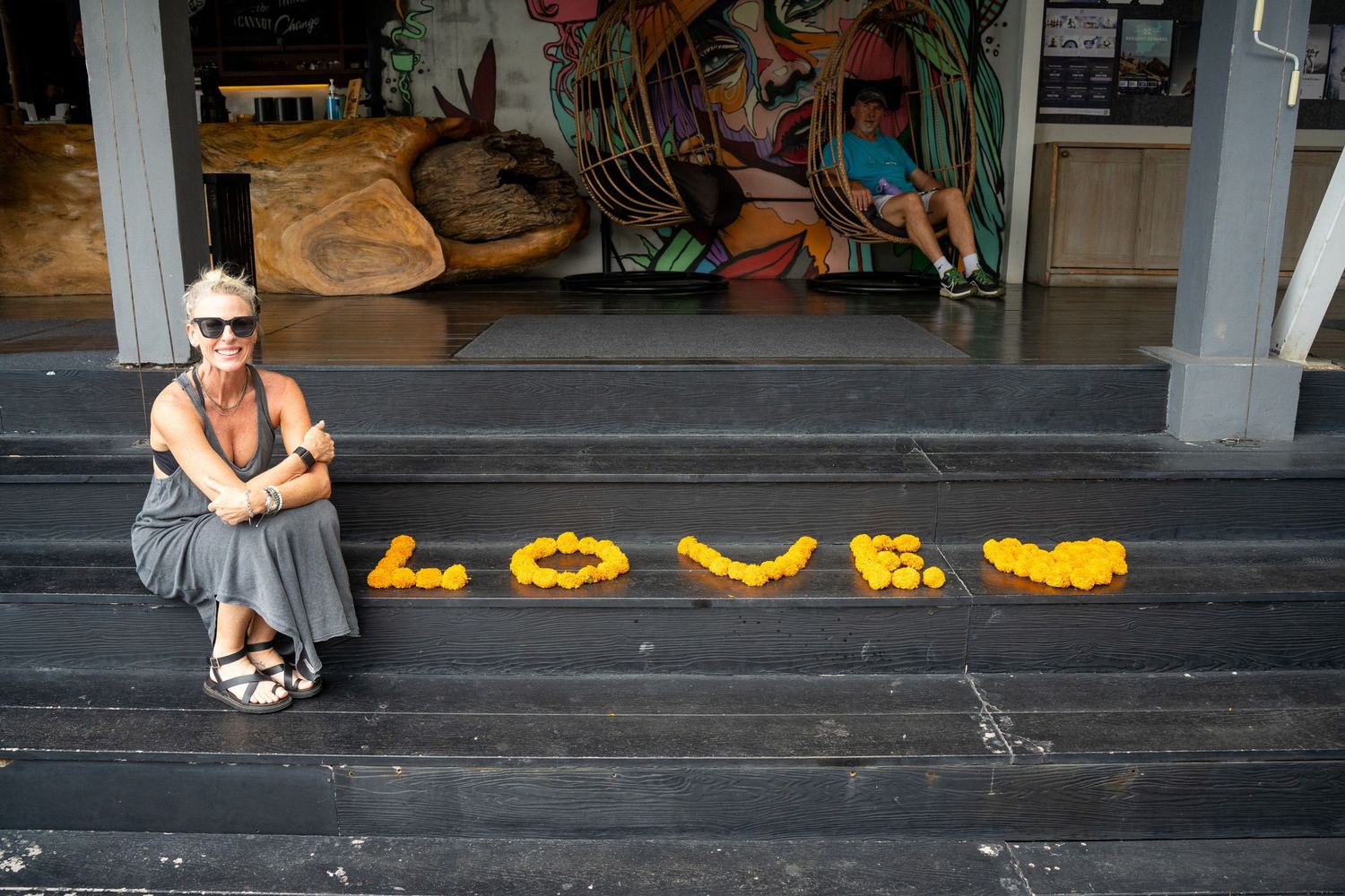 Woman sitting on steps beside yellow flower petals spelling LOVE, with colorful street art mural in background.
