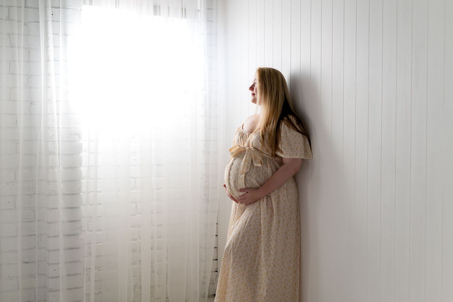 Pregnant woman in beige dress cradling baby bump, standing by white curtain in soft natural light.