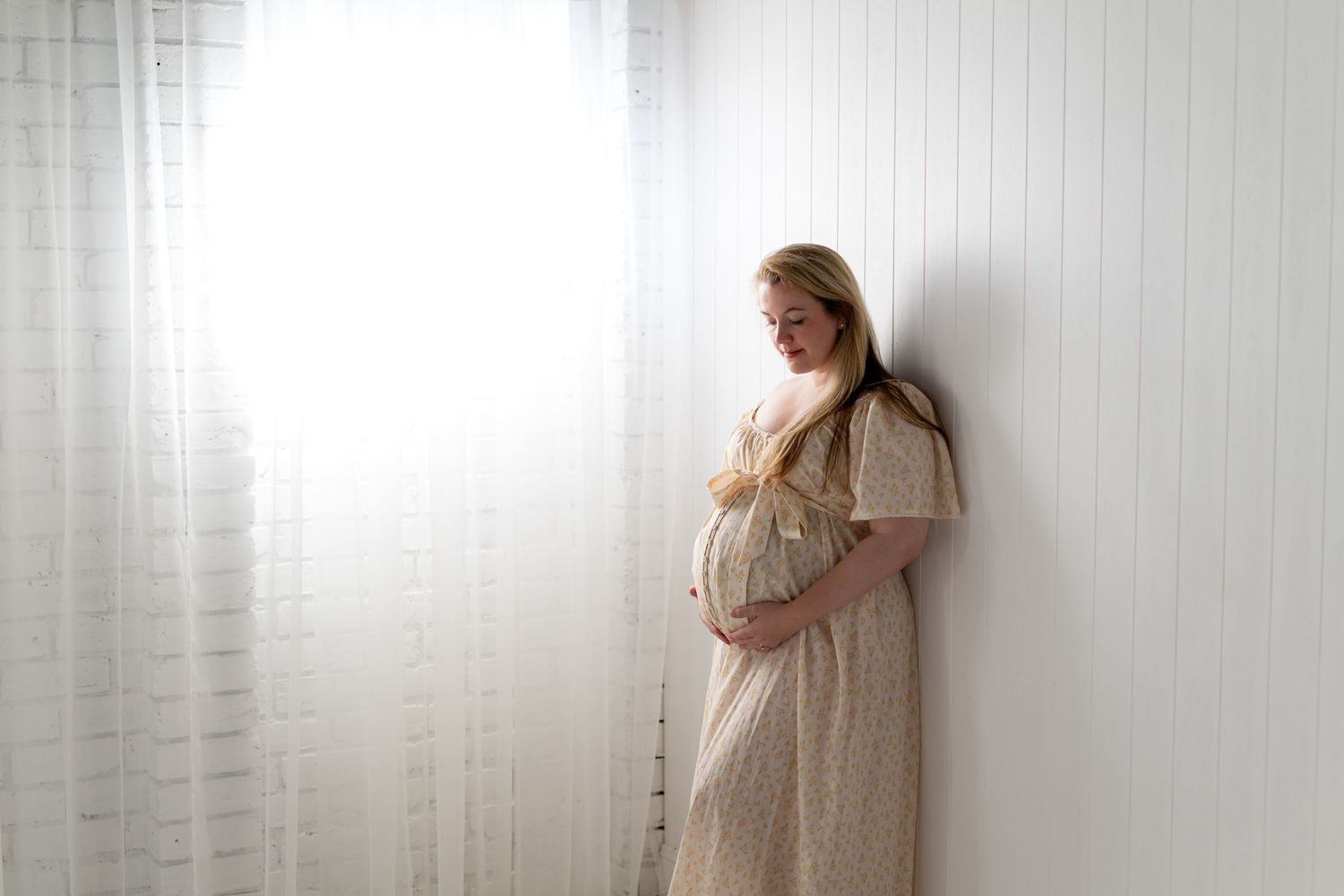Pregnant woman in a cream lace dress cradling her baby bump, leaning against a white wall near sheer curtains.
