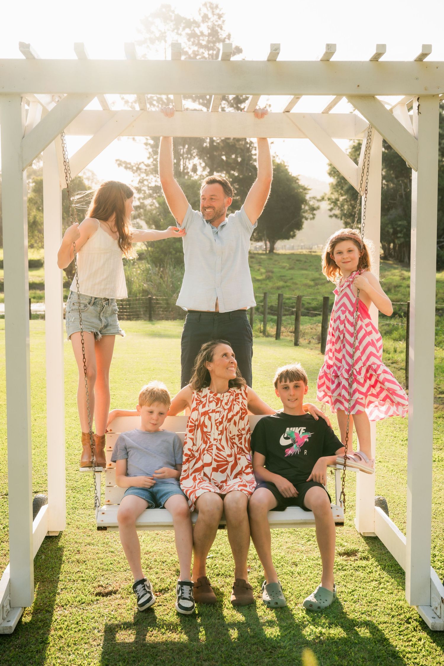 Smiling family of six poses on a white wooden swing in a sunny grassy yard, with parents and four children laughing together.