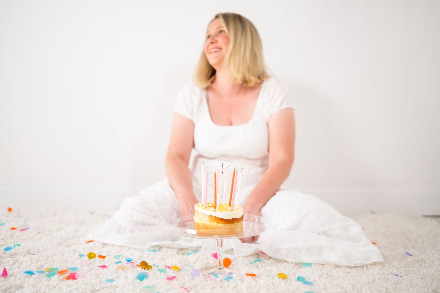 Smiling woman in a white dress sits on a white floor holding a small cake with lit candles, surrounded by scattered colorful confetti.