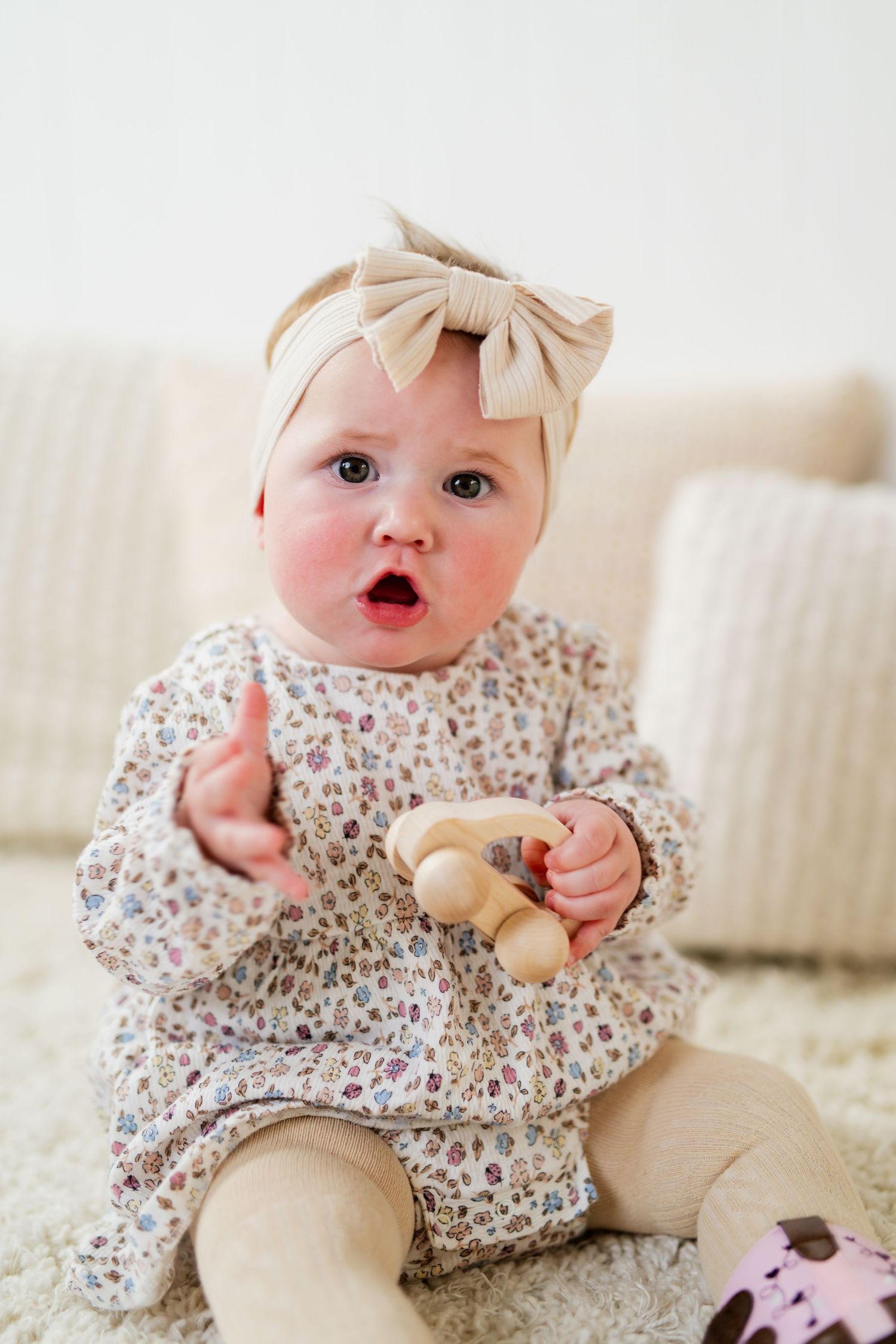 Adorable baby girl in floral dress and bow headband sits on carpet holding wooden toy with mouth open.
