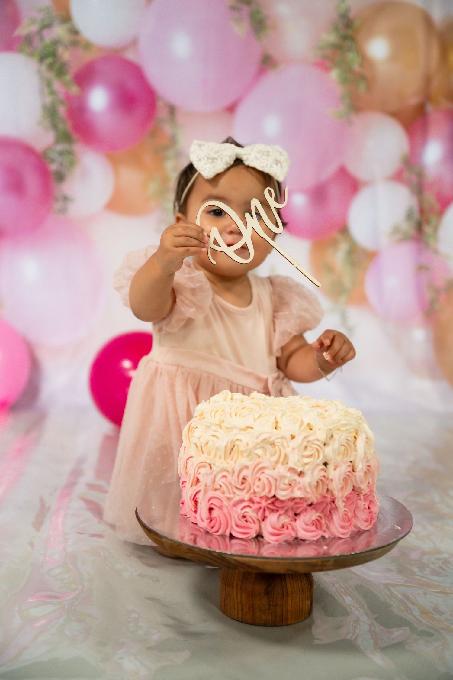 Baby girl in pink dress holds 'One' sign beside a rosette birthday cake, surrounded by pink balloons.