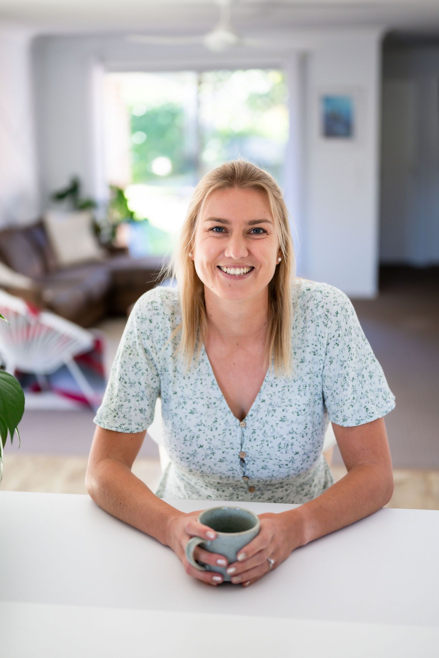 Person in a white floral dress holding a blue ceramic mug while leaning on a white counter in a bright interior space.
