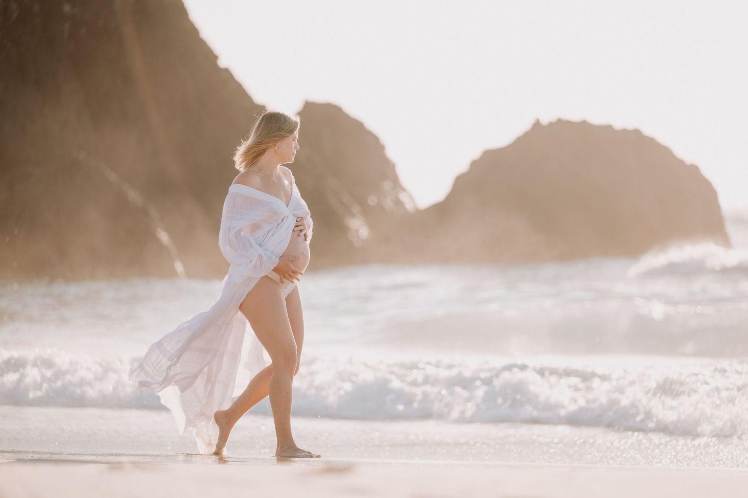 Pregnant woman in flowing white dress walking on a sunlit beach with rocky cliffs and ocean waves.