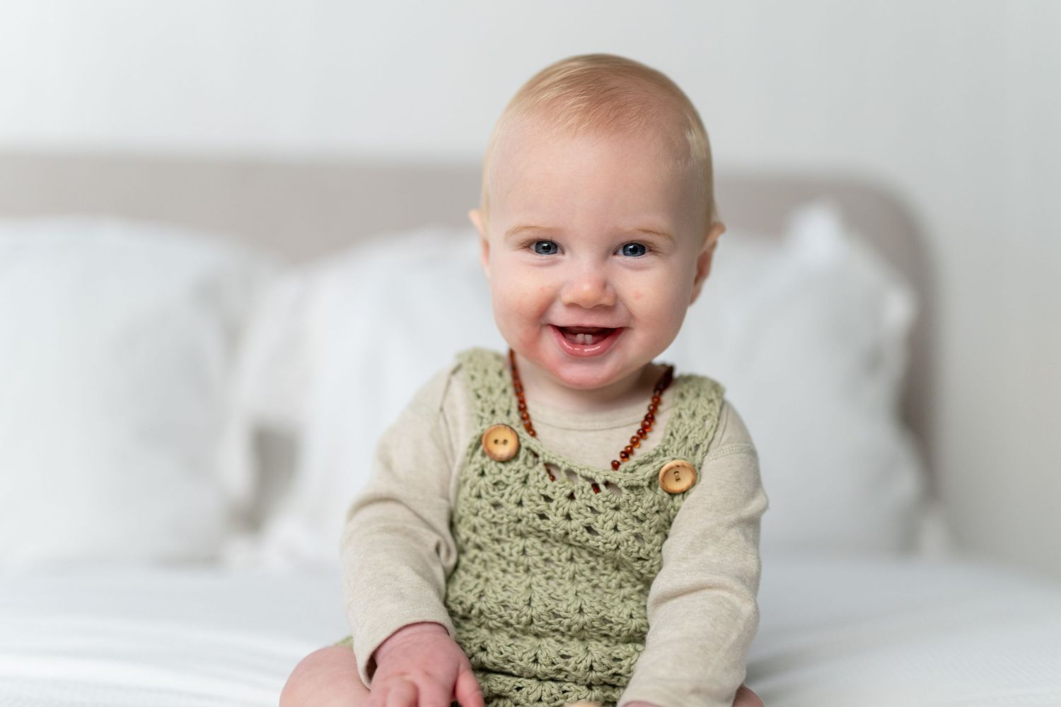 Smiling baby in green crochet overalls and cream shirt sitting on white bed, happy infant portrait.
