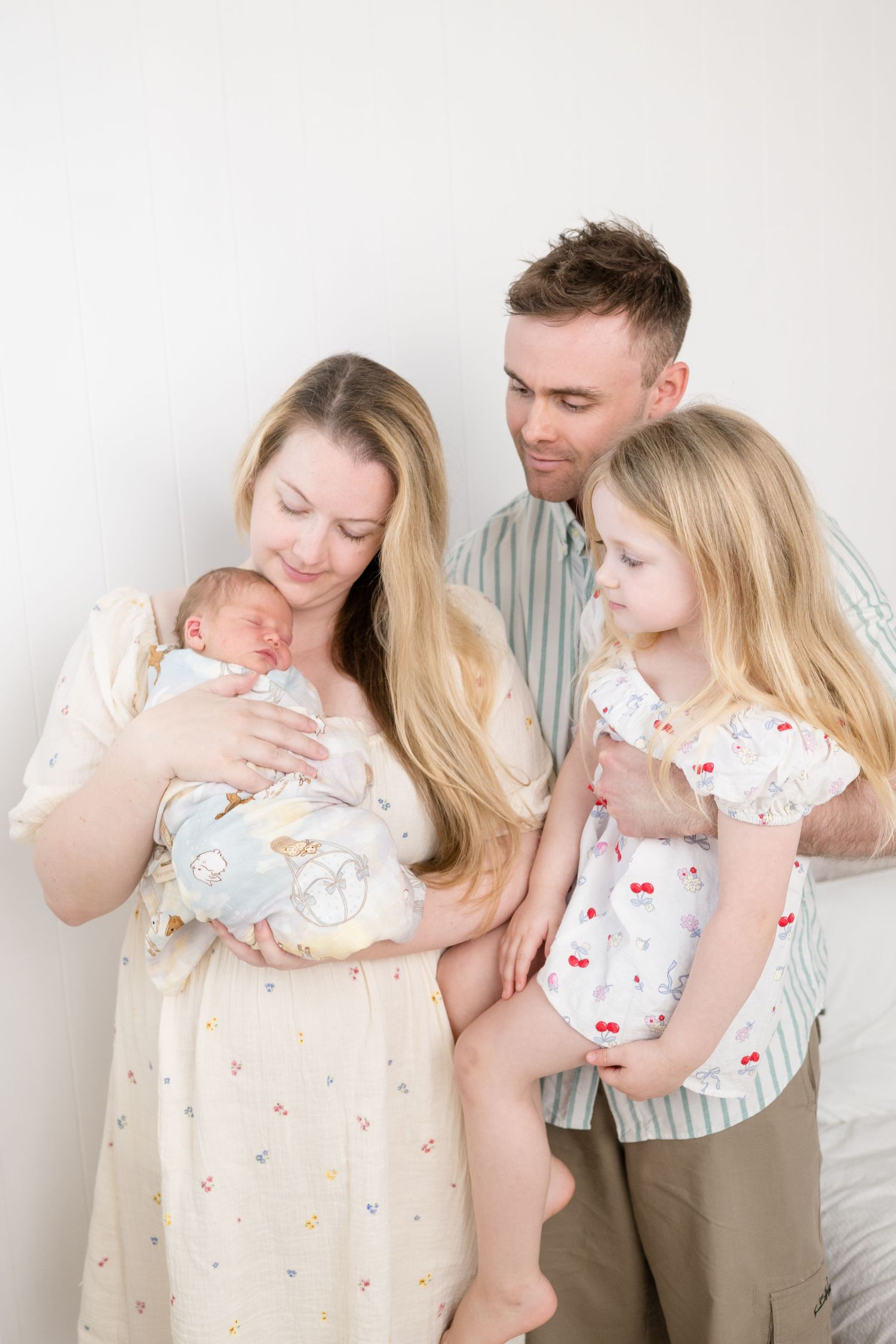 A happy family of four with parents holding a newborn baby and young blonde girl in a bright white studio setting.