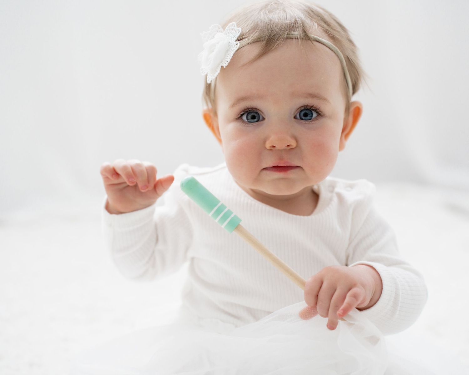 Adorable baby in white outfit holding a colorful toothbrush against a soft white background.