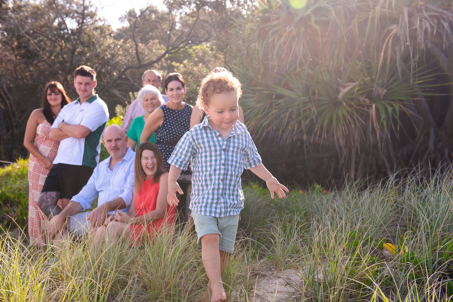 A young boy runs barefoot on sandy path while multi-generational family poses together among lush green foliage outdoors.