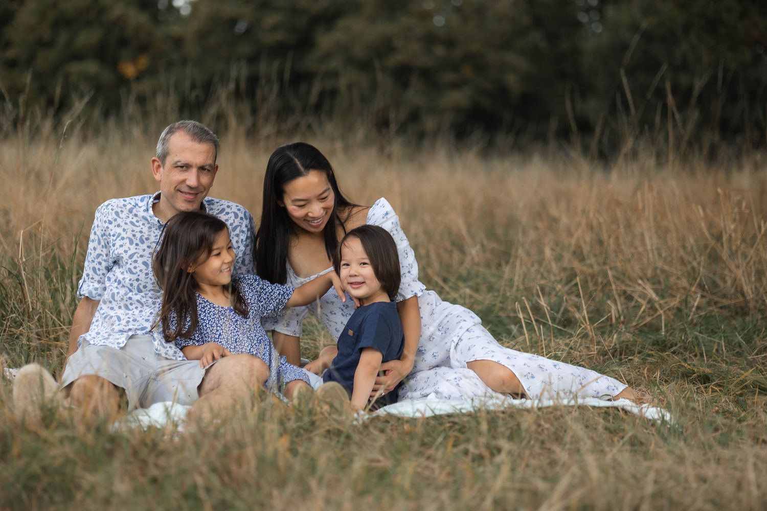 Family on a summer photo shoot, dressed in similar shades of blue and white, sat on a picnic blanket smiling together.