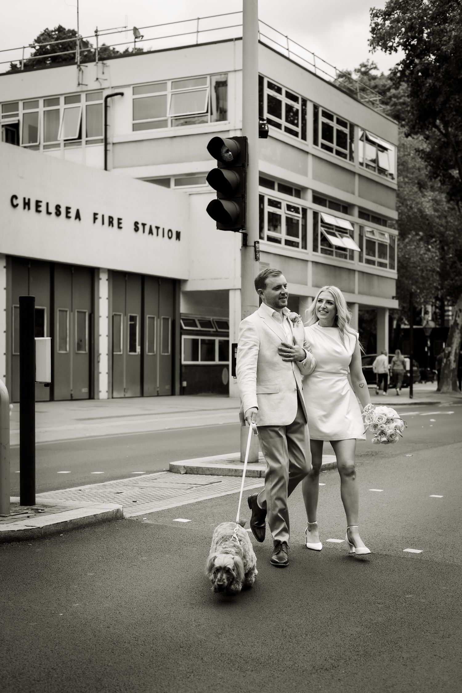 Relaxed image of bride and groom crossing the road in London, with Chelsea Fire Station in the background.