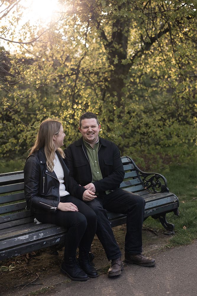 Relaxed couple photoshoot, with couples sat on a bench laughing and joking together in London park at golden hour.
