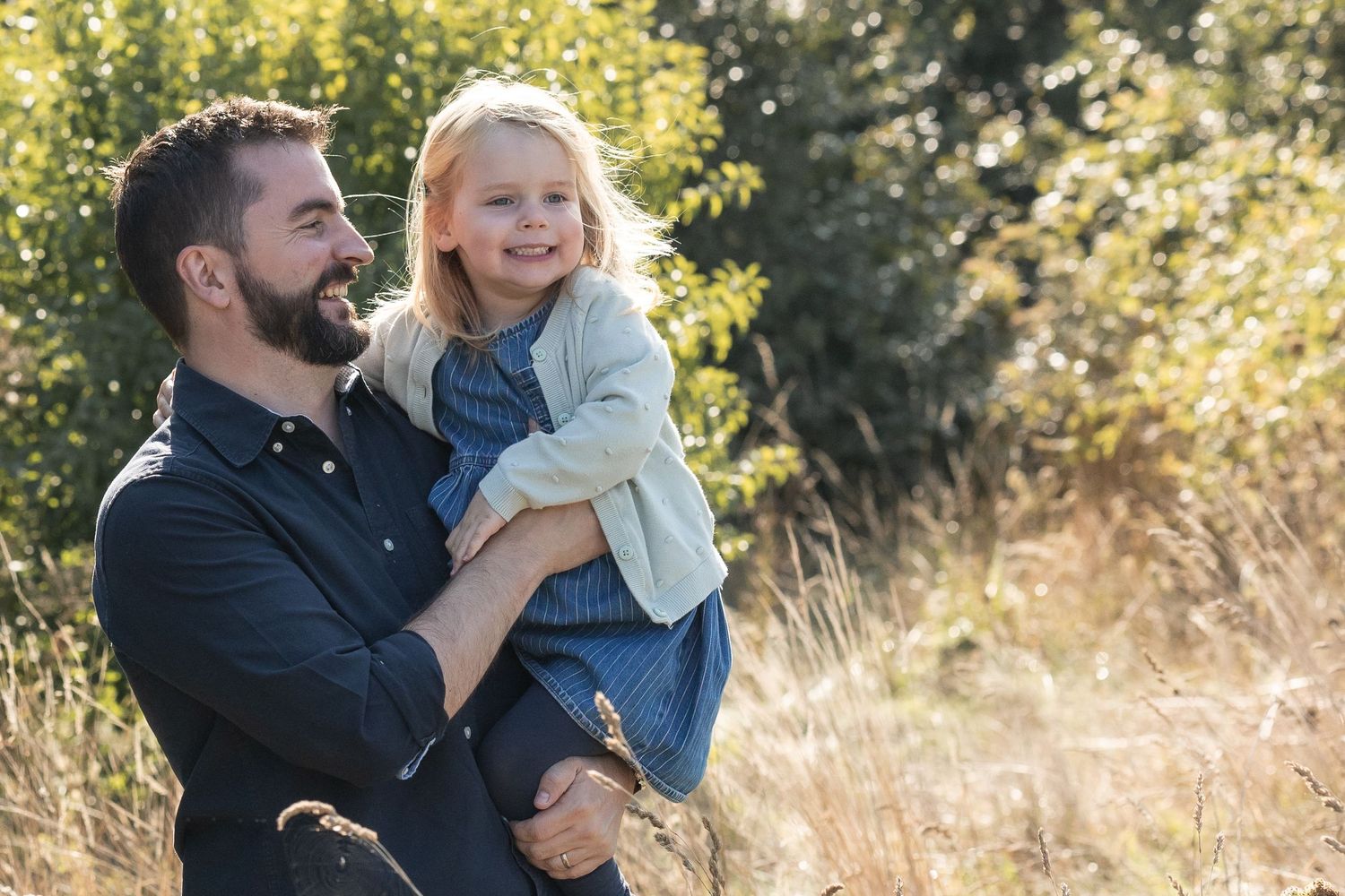 Image of a father carrying his daughter and she is smiling. The sun has caught her hair, and the blonde hair is glowing.