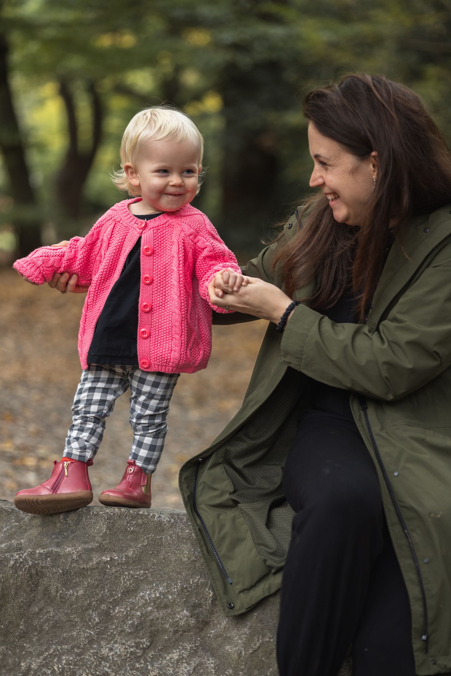 Toddler standing on a tree stump holding mum's hand whilst they look at each other lovingly.
