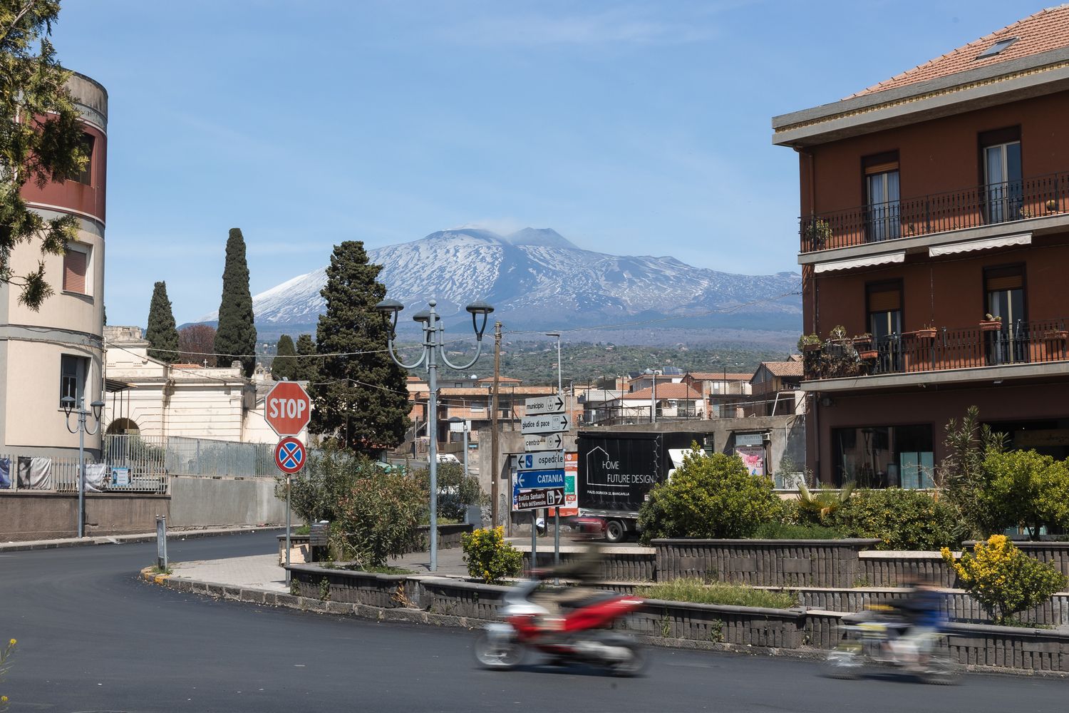 Landscape image of a roundabout, with Etna in the background. Slow shutter speed blurs two motorcycles in the foreground of the image.