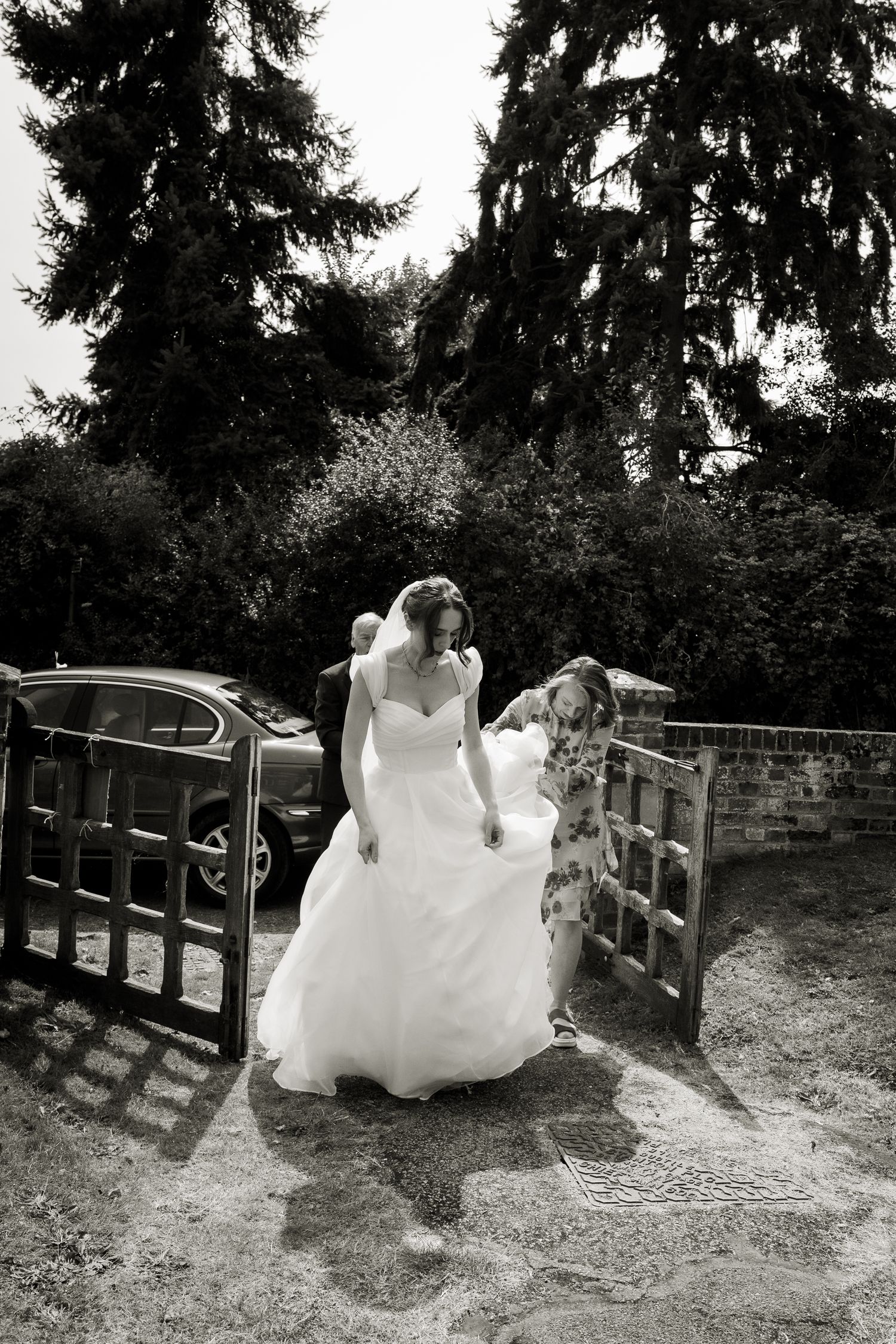 Black and white image of the bride arriving at the church. Bridesmaids are holding the train of her dress which is glowing white in the sun.