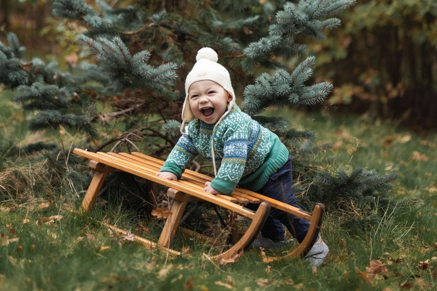 Young boy grinning on an outdoor Christmas shoot. He's wearing a white bobble hat and leaning on a vintage sledge.