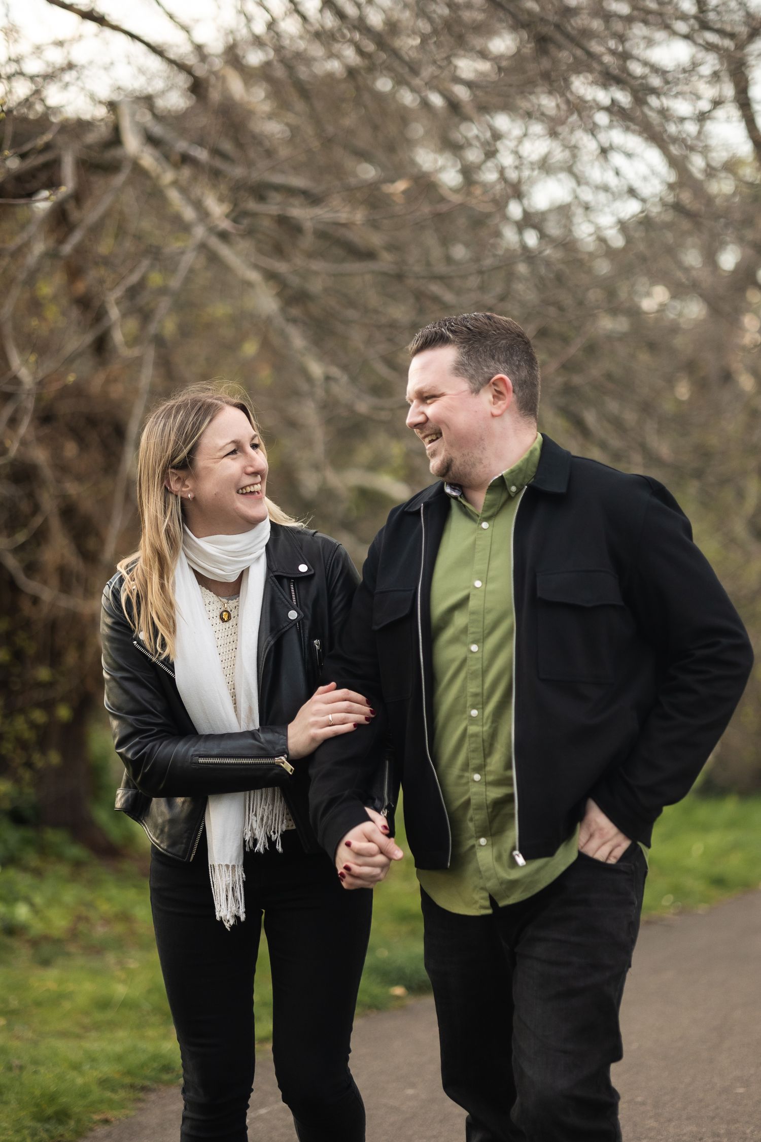 Couple pre-wedding photo shoot in Greenwich park, London. They are strolling hand in hand, smiling at each other.