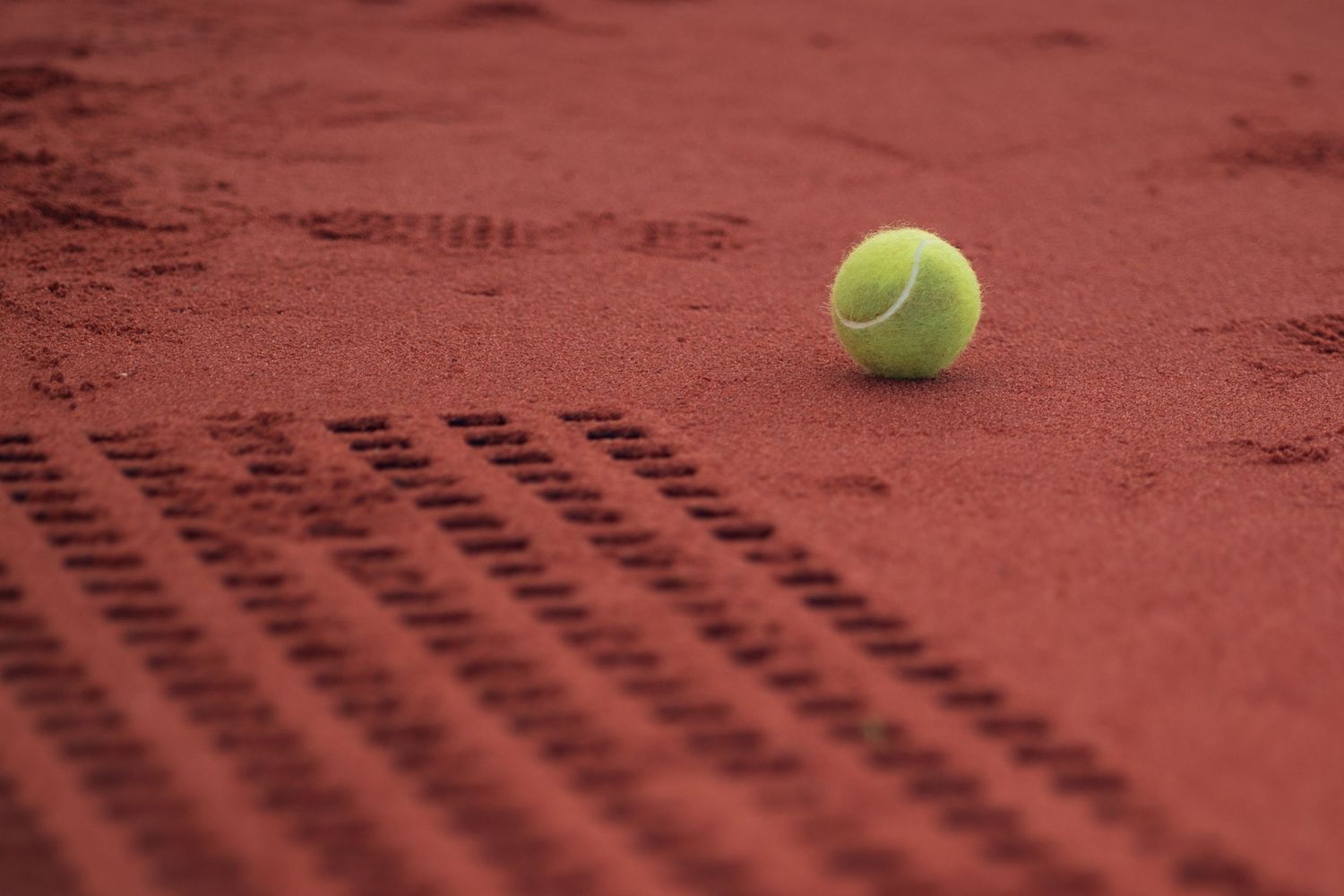 A lone yellow tennis ball on orange clay court.