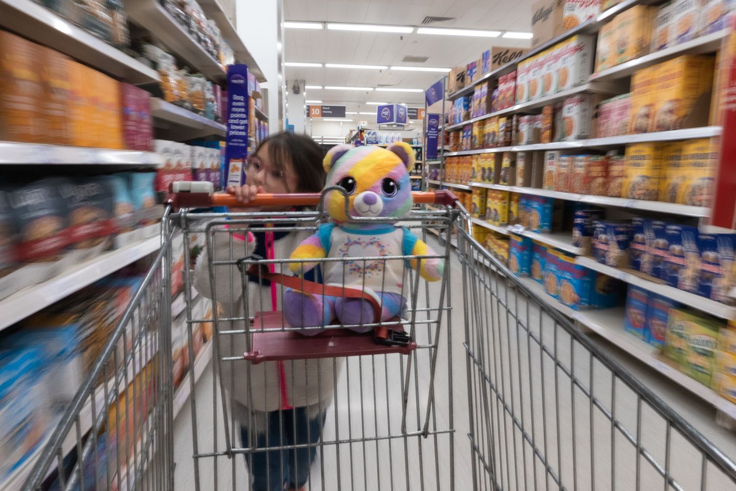 Camera attached to the supermarket trolley, slow shutter, focussing in on teddy sitting in the seat, whilst being pushed by young girl.