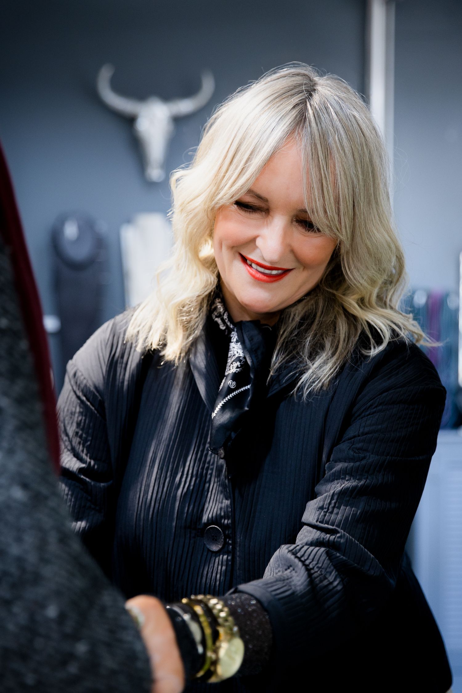 Smiling blonde woman in black jacket with bracelets, standing in a boutique with a bull skull decor.