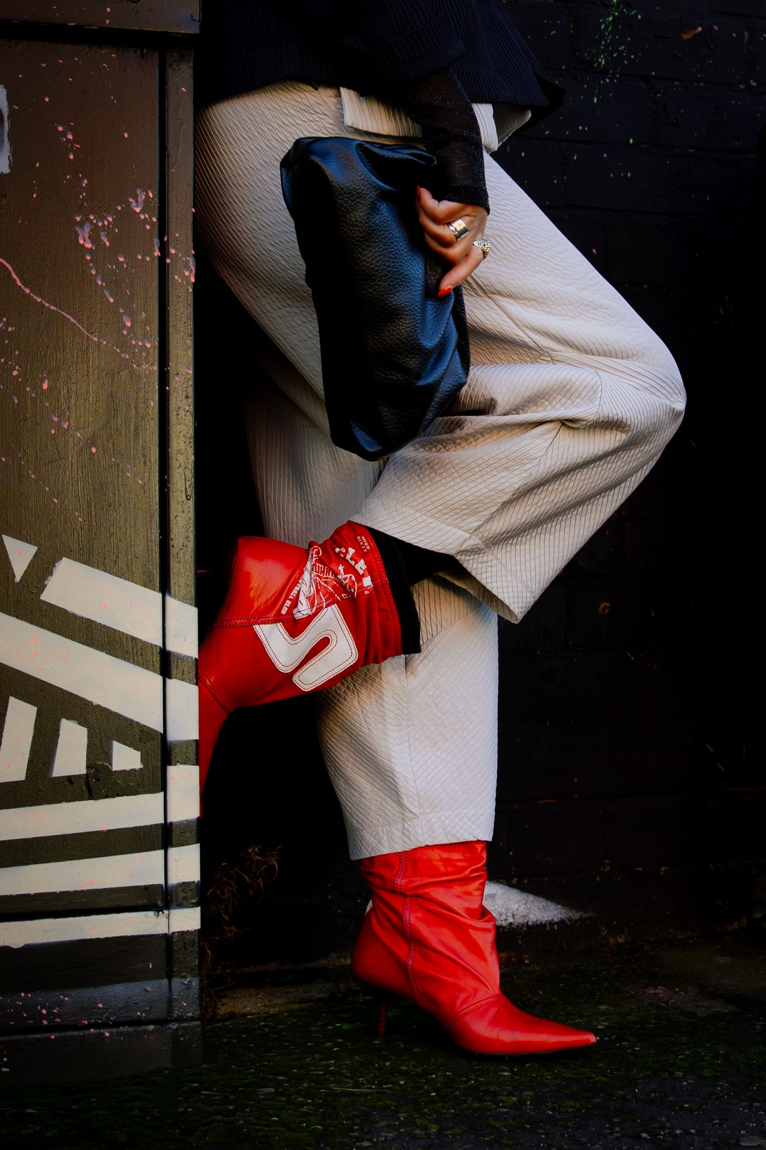 Person wearing cream pinstripe trousers and bold red pointed-toe ankle boots with white logo detail, leaning against dark wall.