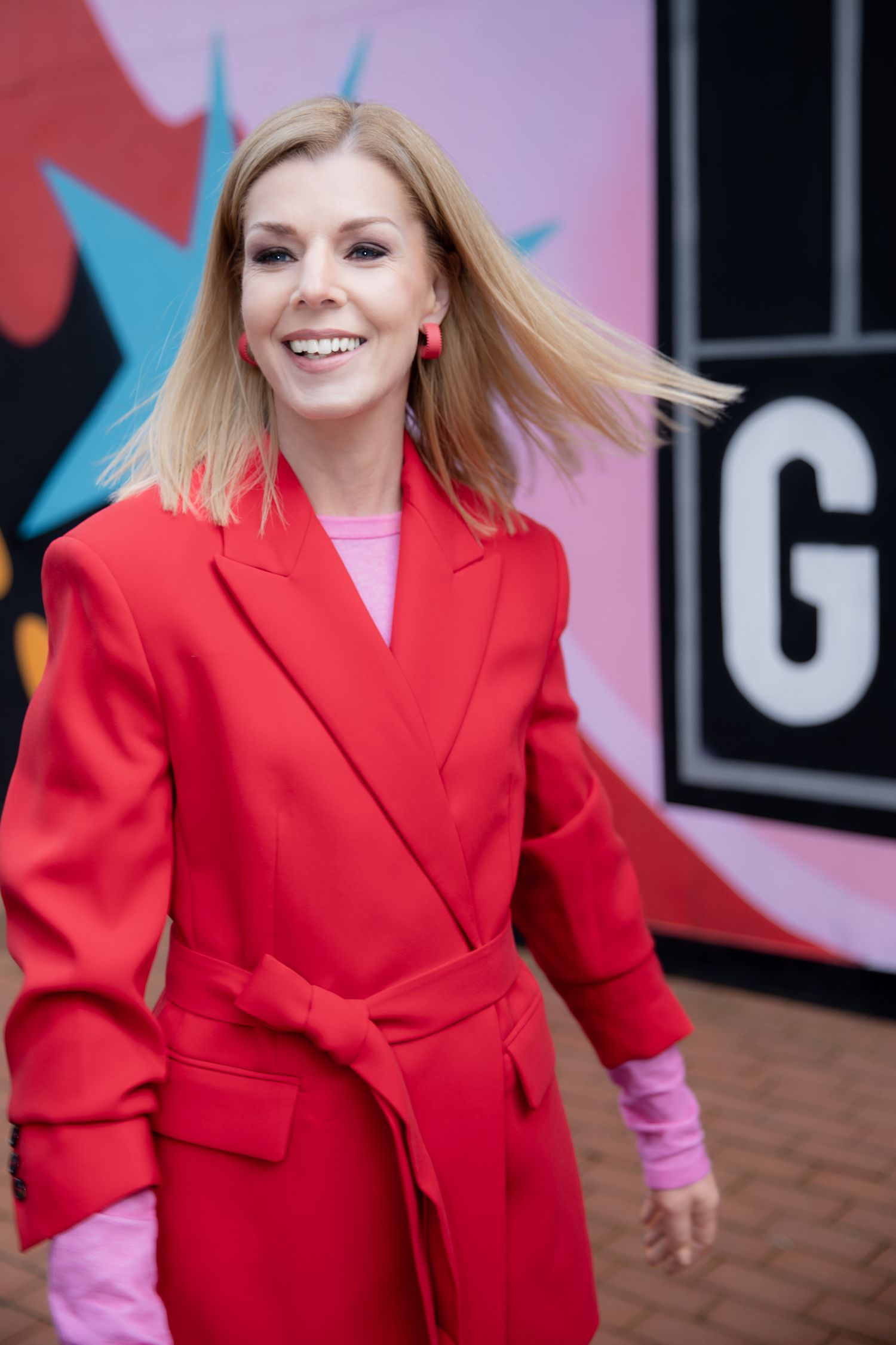 Smiling blonde woman in a red belted coat and pink sweater posing outdoors in front of a colorful mural.