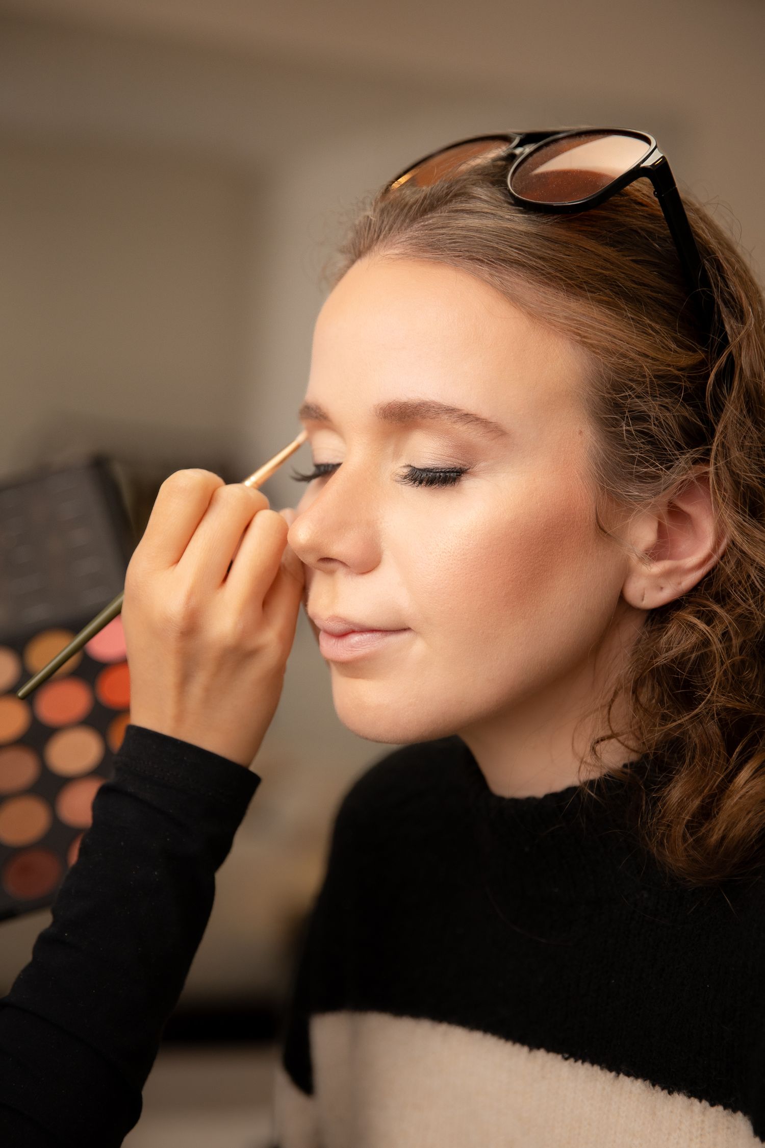 Makeup artist applying eyeshadow to a young woman with sunglasses on her head, near a colorful palette.