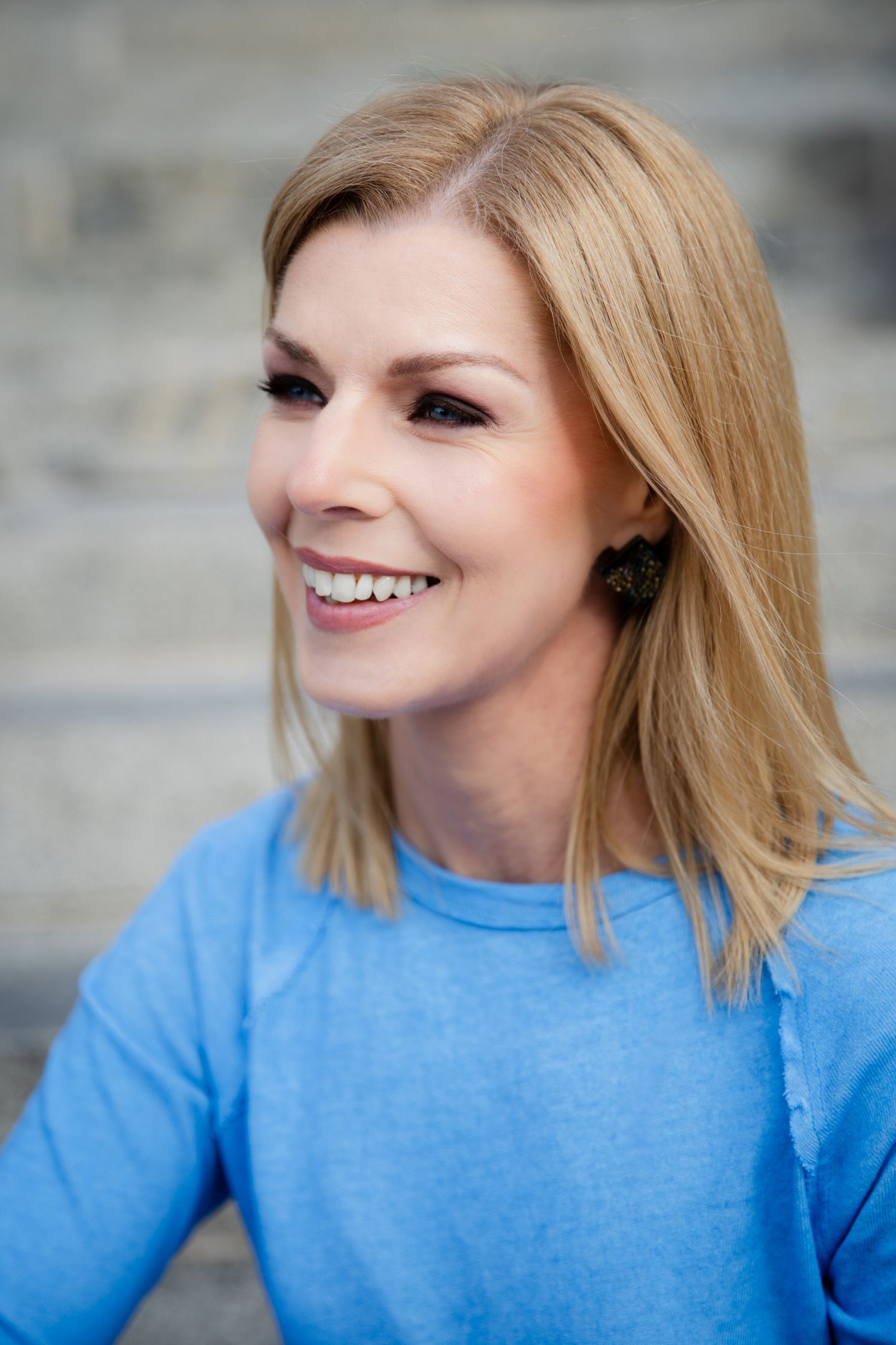 Smiling blonde woman in blue t-shirt with shoulder-length hair and earrings outdoors.