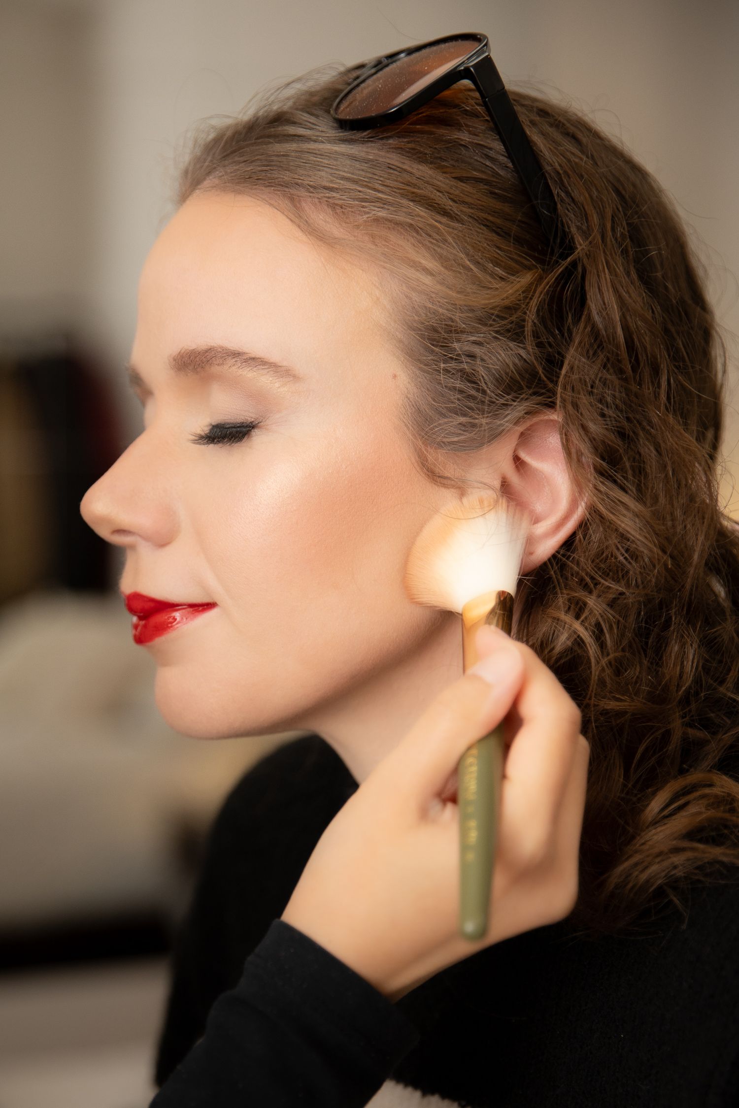 A makeup artist applies blush to a young woman with red lips and sunglasses on her head using a brush.