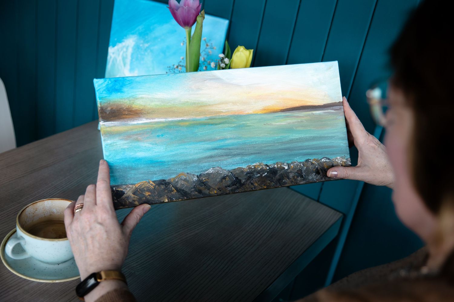 Woman holding a small coastal landscape painting with textured rocky edge, coffee cup and flowers visible in background.