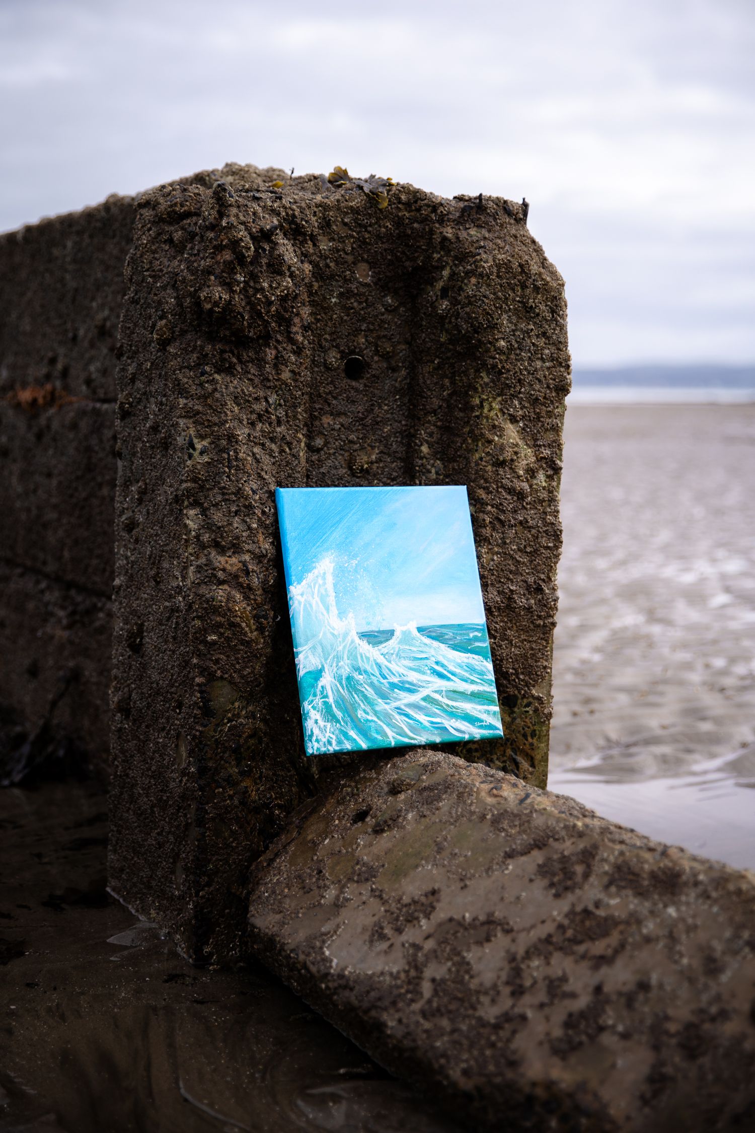A vibrant ocean wave painting leaning against a weathered stone groyne on a sandy beach.