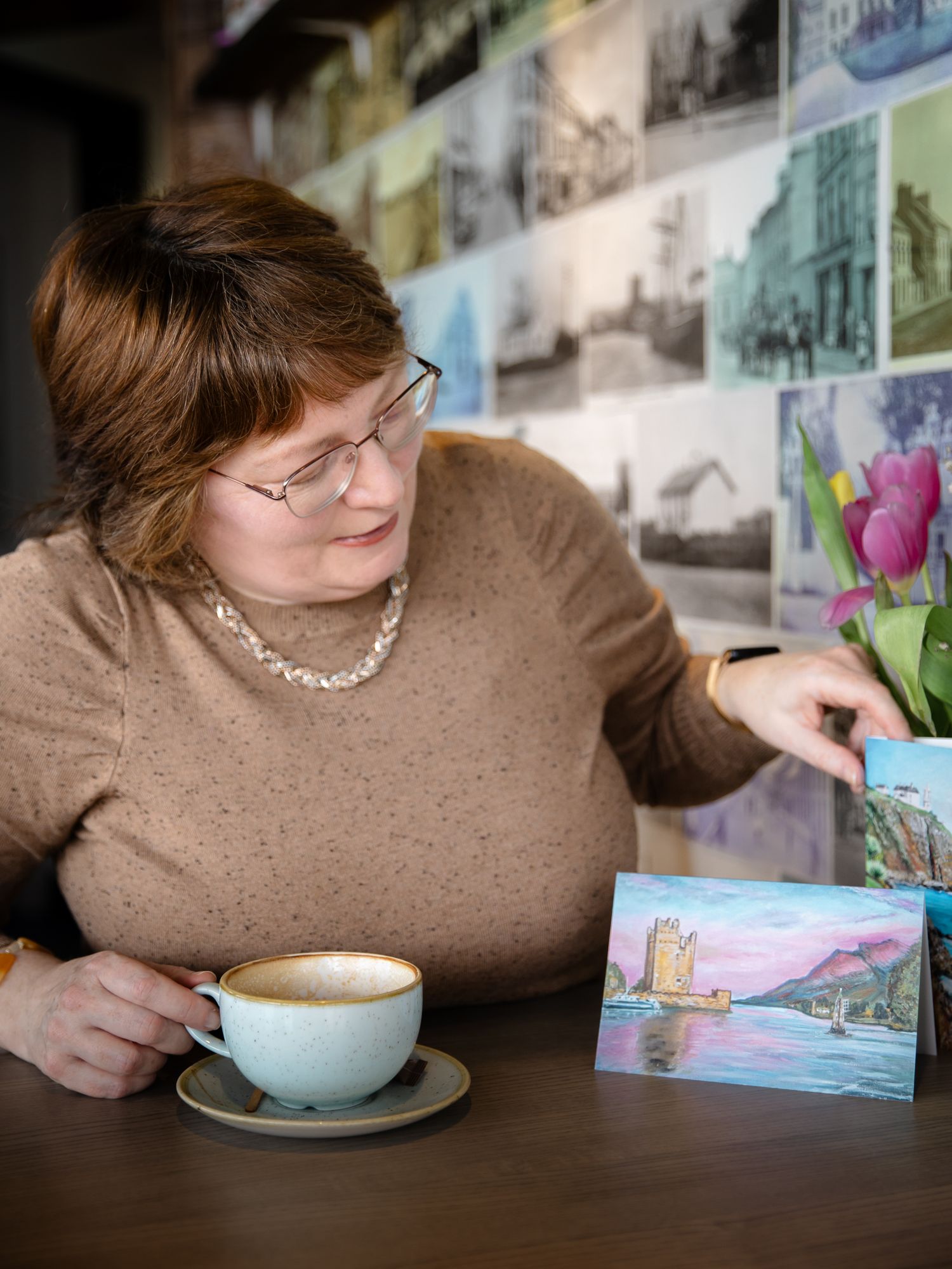 Woman with glasses enjoying coffee at cafe table, examining colorful landscape painting card beside pink tulips.