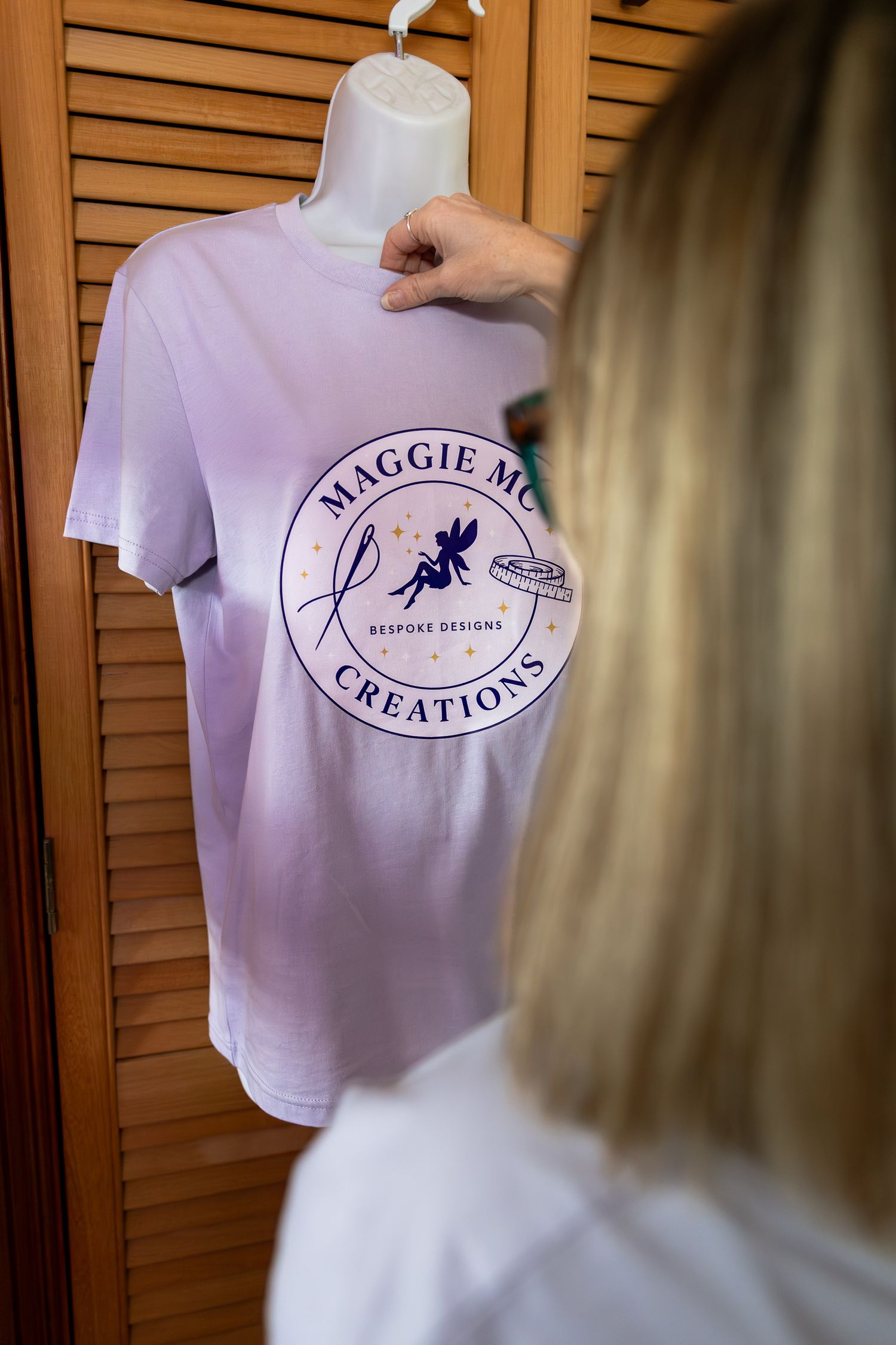 A lavender t-shirt with Maggie Mo Creations bespoke designs logo displayed on a mannequin against wooden shutters.