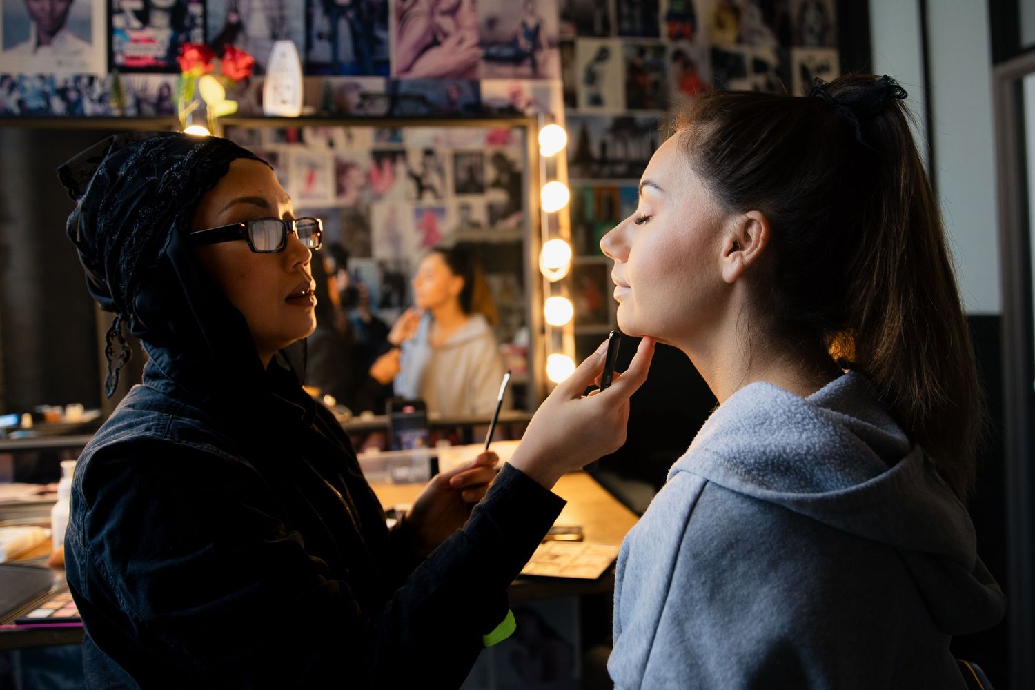 Makeup artist applying foundation to a young woman in a professional vanity-lit dressing room.