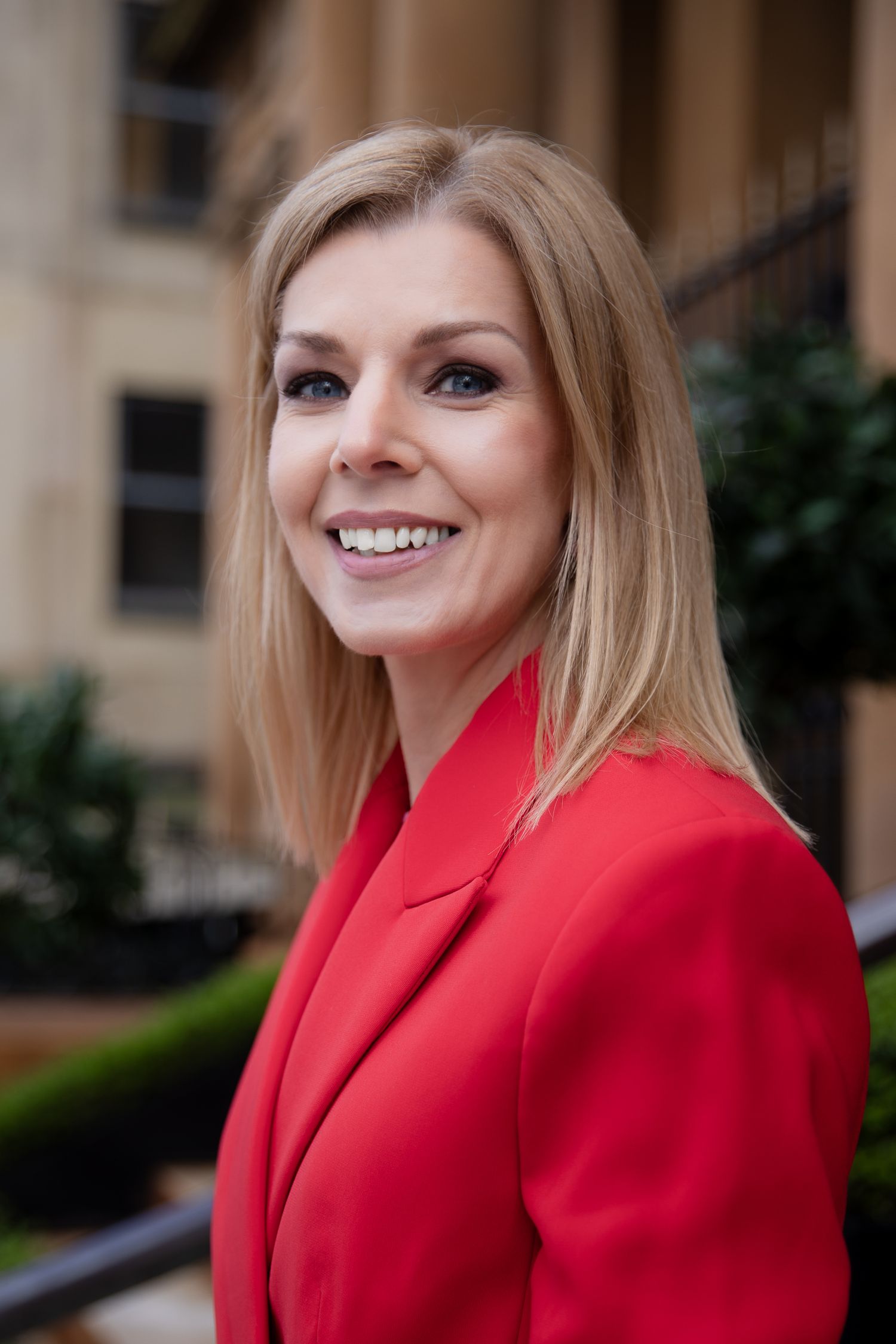 Professional blonde woman smiling in a red blazer standing outdoors near a classic stone building.