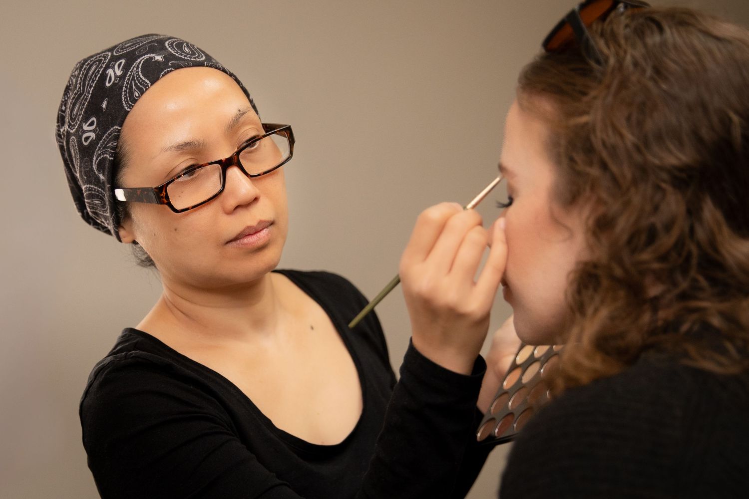 Makeup artist applying eyeshadow to a woman, wearing glasses and a black headscarf, in a studio setting.
