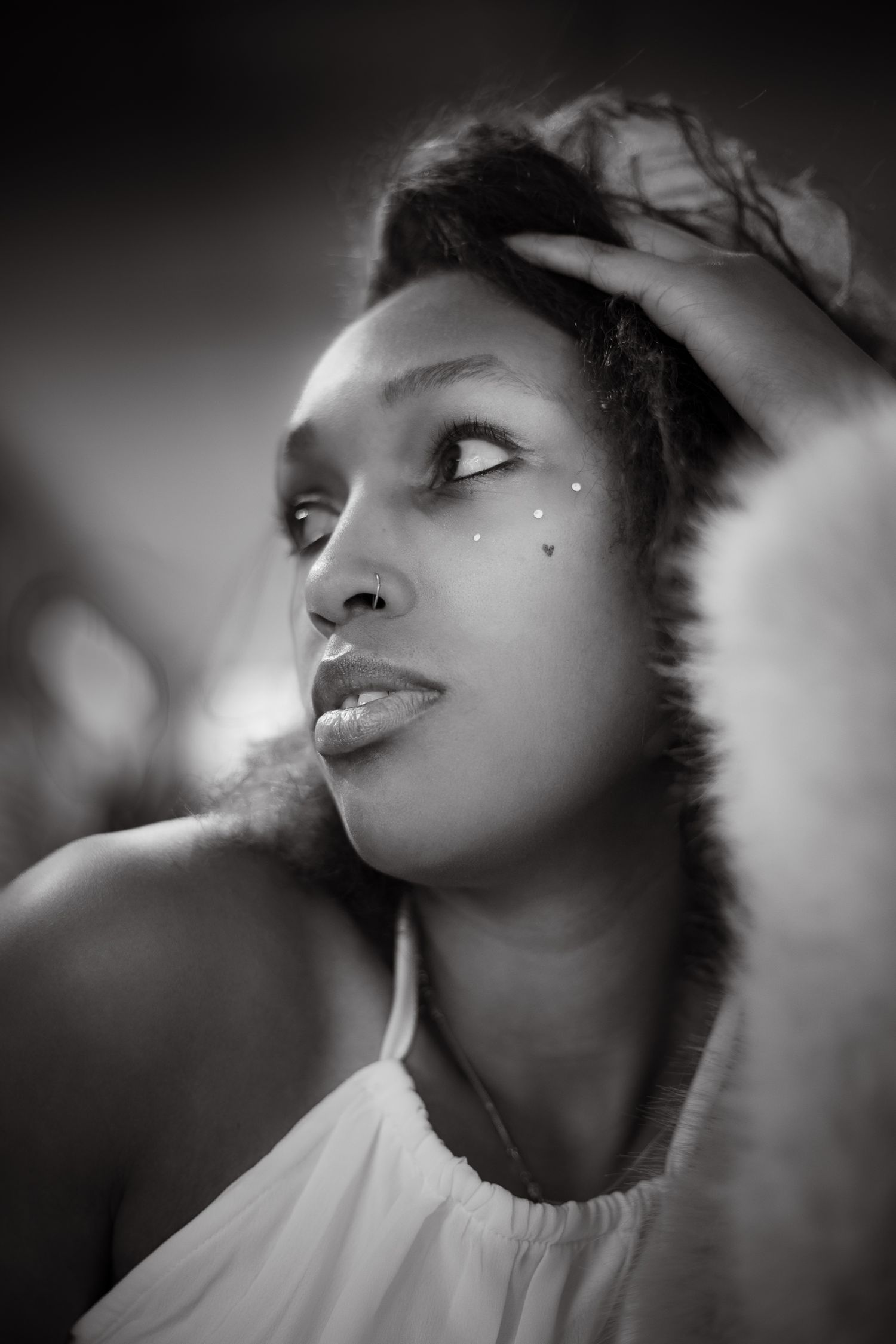 Black and white portrait of a young woman with face gems, hand in hair, wearing white top, gazing upward.