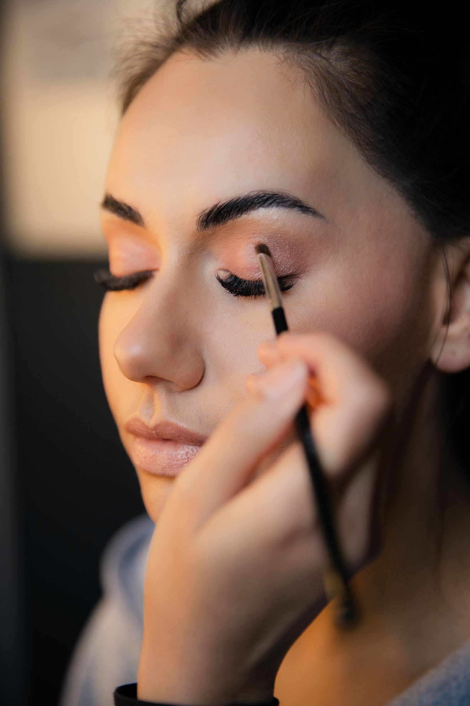 Professional makeup artist applying eyeshadow with brush on young woman with dark hair in beauty salon.