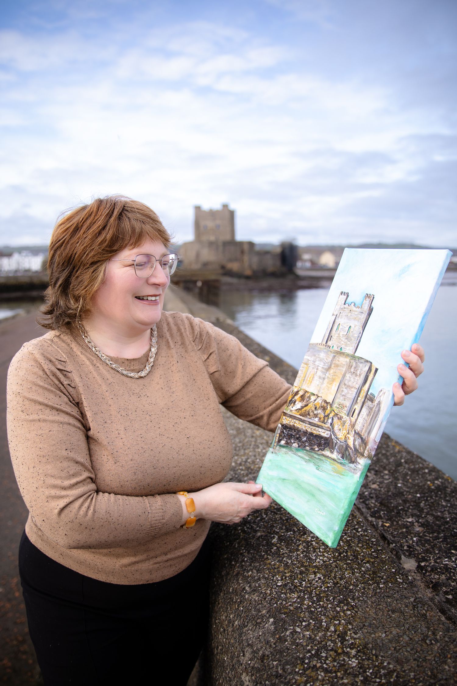 Woman holding a painting of a castle by the waterfront with a historic stone castle visible in the background.