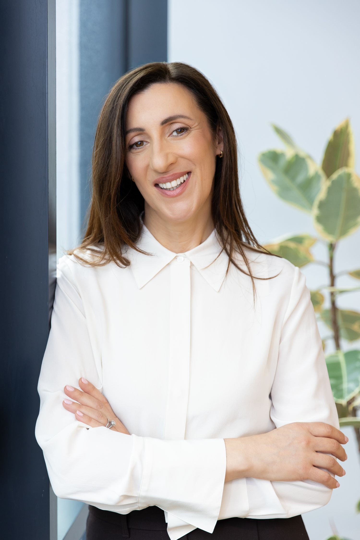 Professional woman in white blouse smiling with arms crossed in modern office setting.