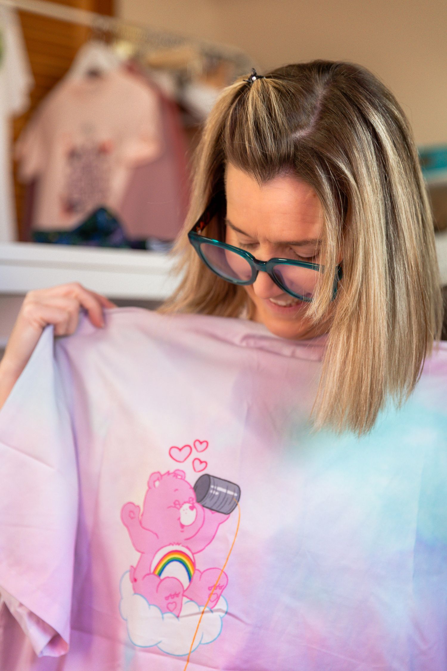 Woman with glasses holding a pink tie-dye Care Bears graphic t-shirt in a clothing store.