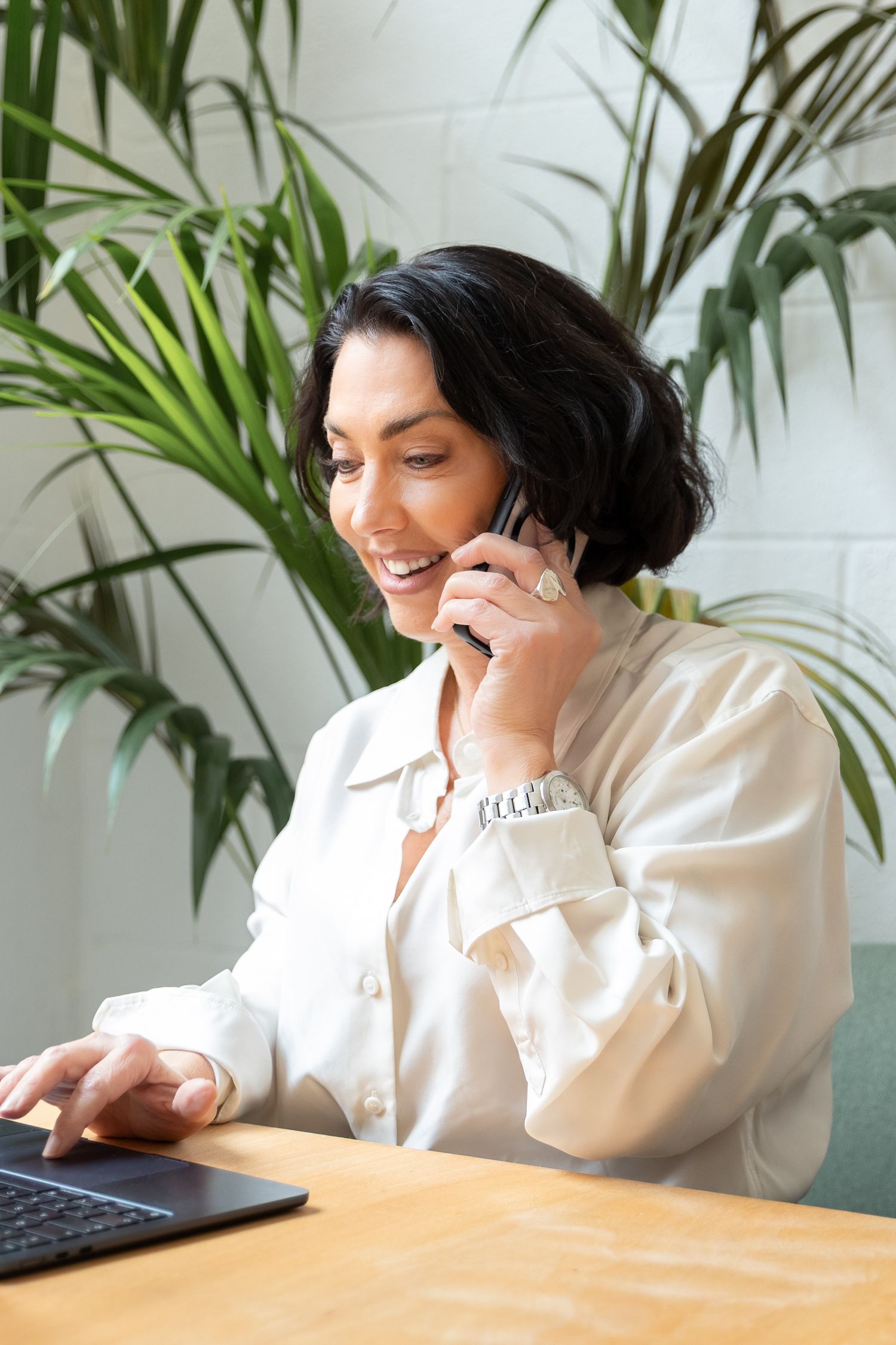 Smiling businesswoman talking on phone while working on laptop at desk with green plants in background.