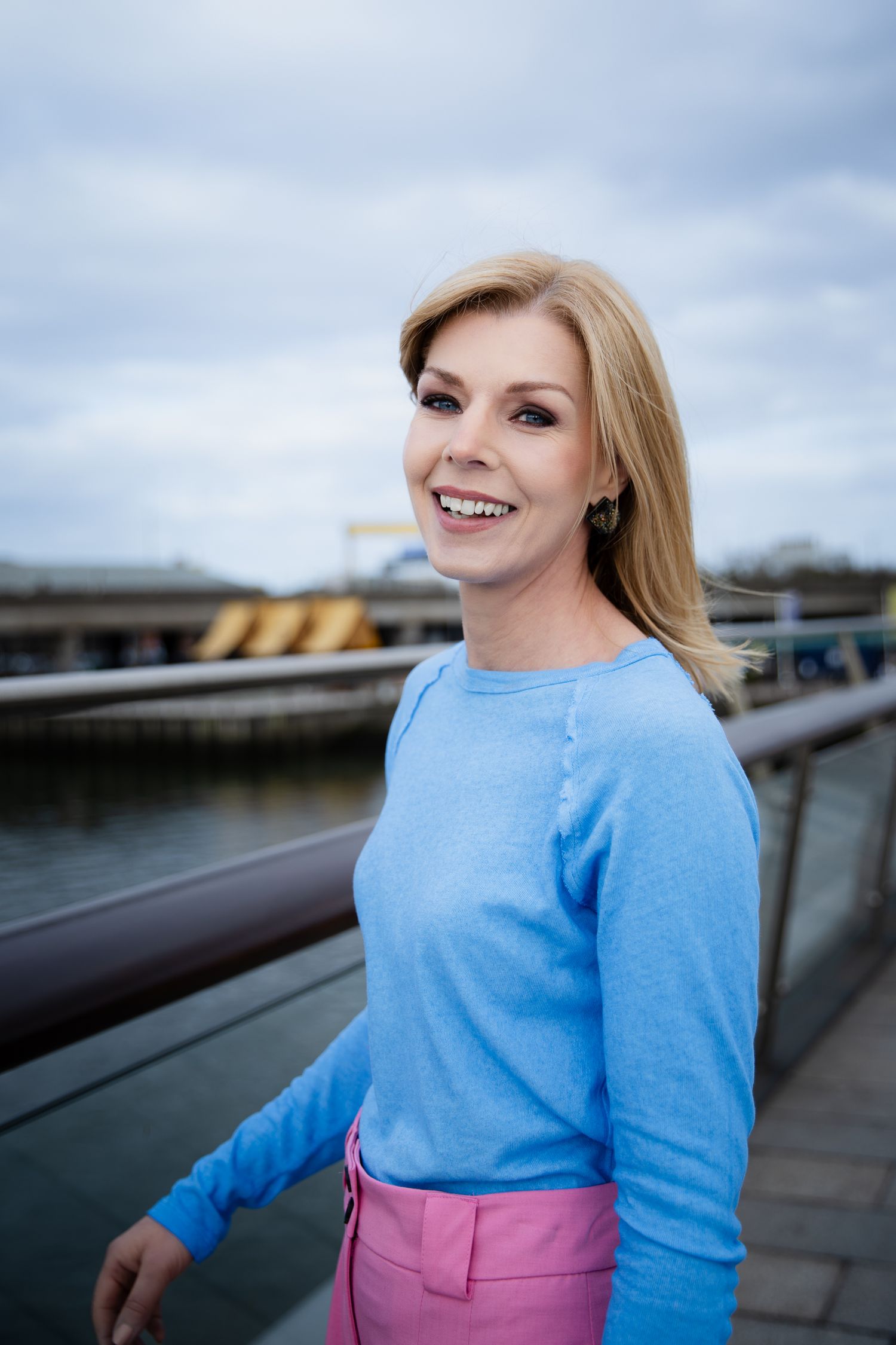 Smiling blonde woman in blue sweater and pink pants standing by waterfront railing on cloudy day.