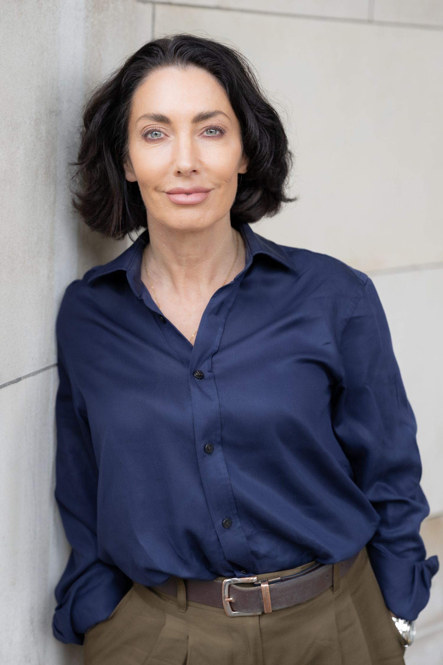 Professional woman in navy blue shirt and olive pants posing confidently against a light stone wall.