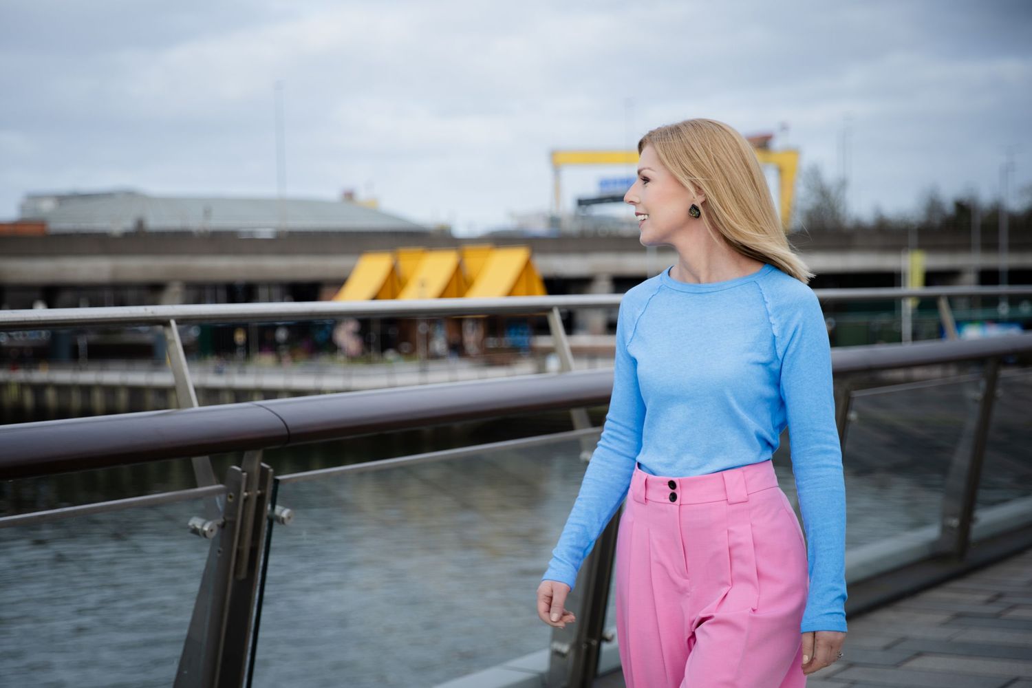 Blonde woman in blue top and pink trousers standing on a waterfront bridge with industrial cranes in the background.