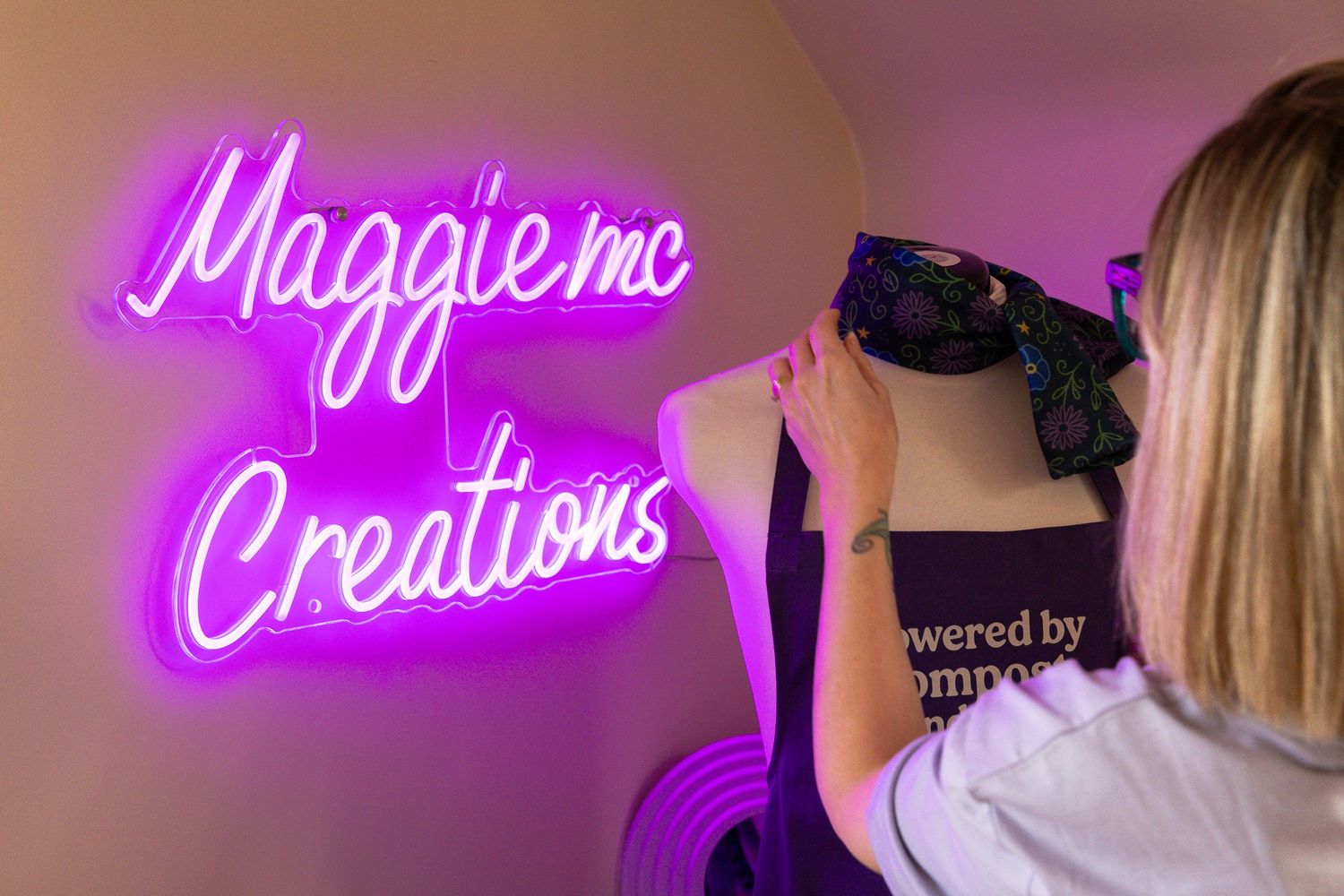Woman arranging display near pink neon sign reading Maggie MC Creations on a wall.