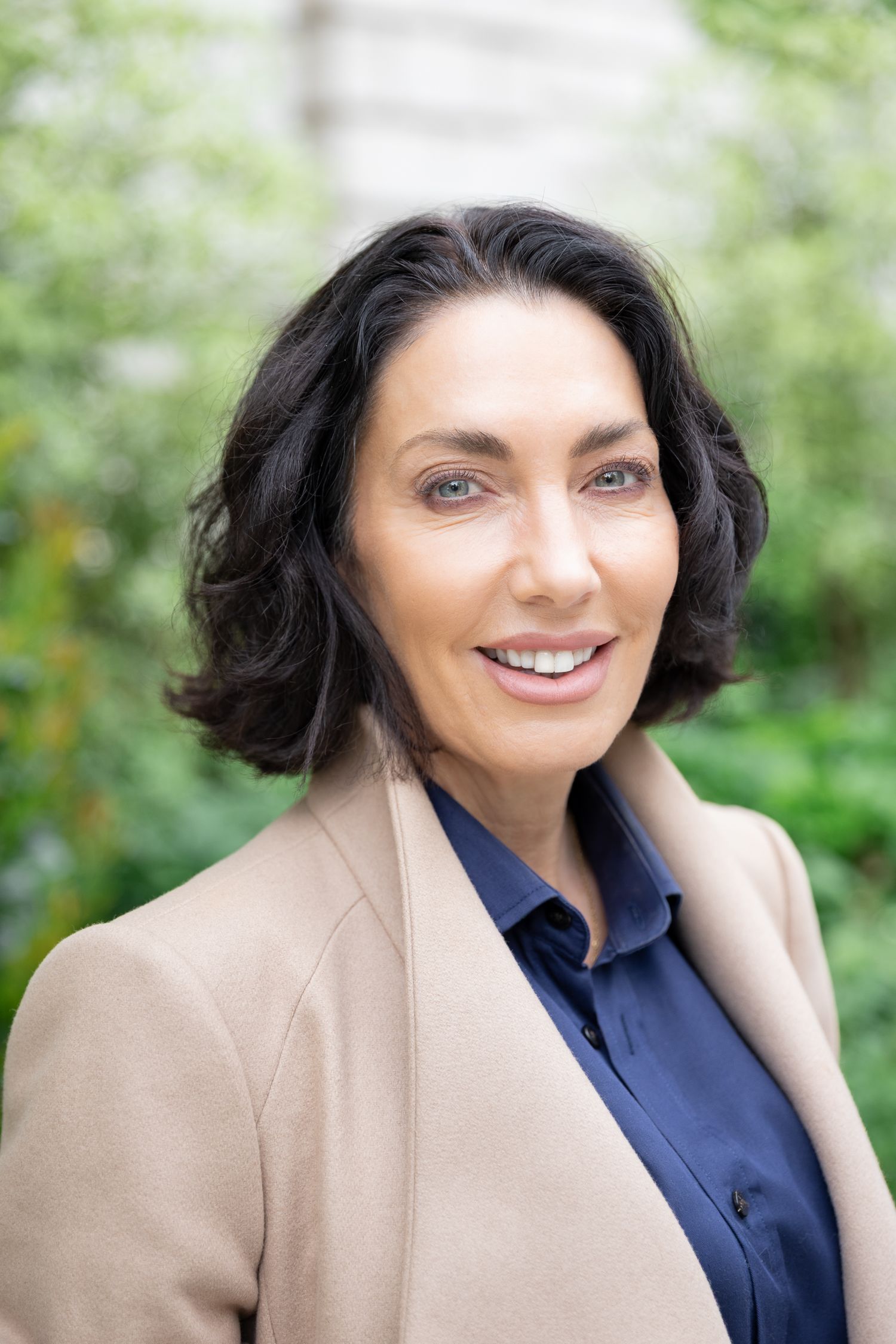 Professional woman with dark hair smiling outdoors in a beige blazer and navy shirt.