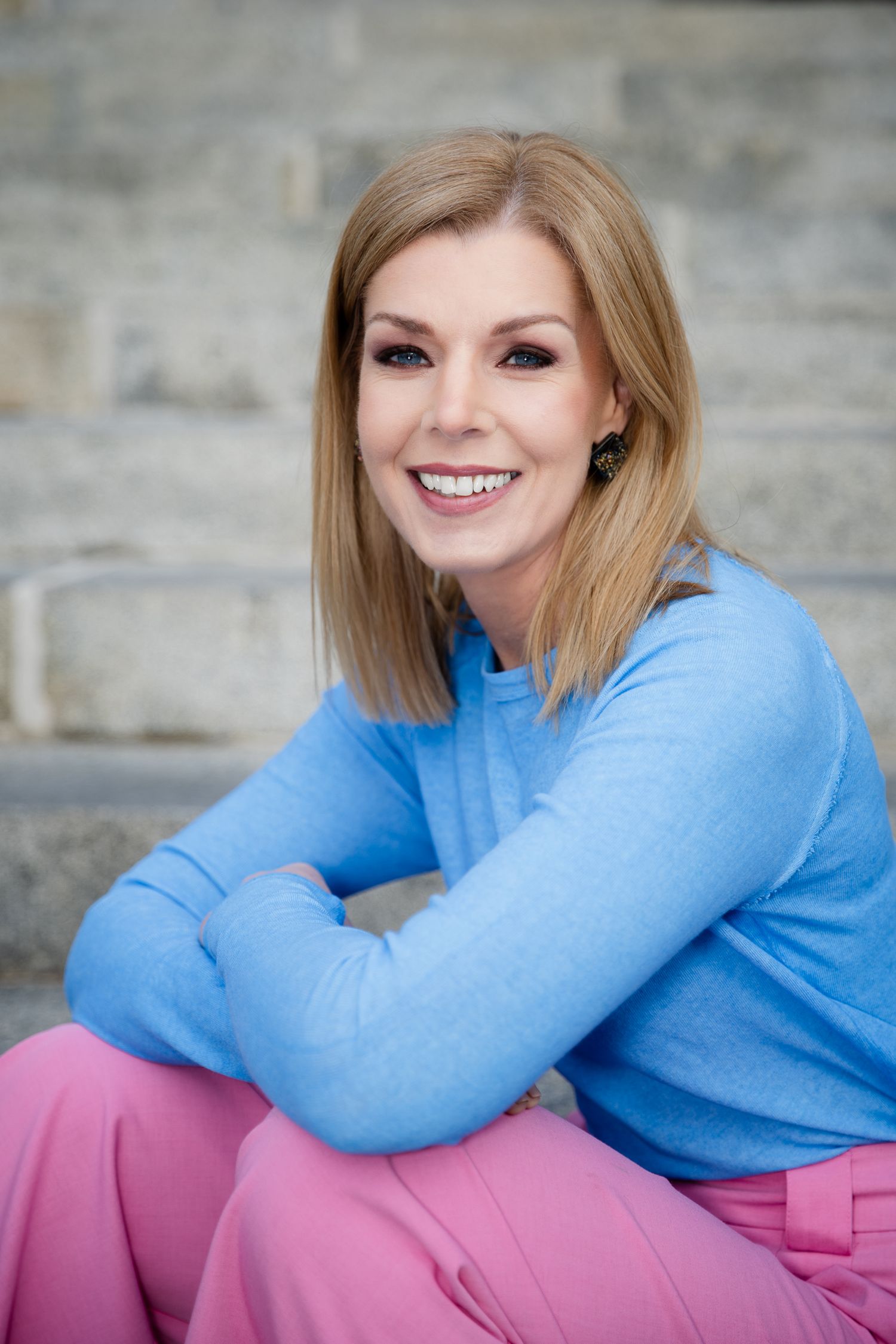 Smiling blonde woman in blue sweater and pink pants sitting on stone steps with arms crossed.