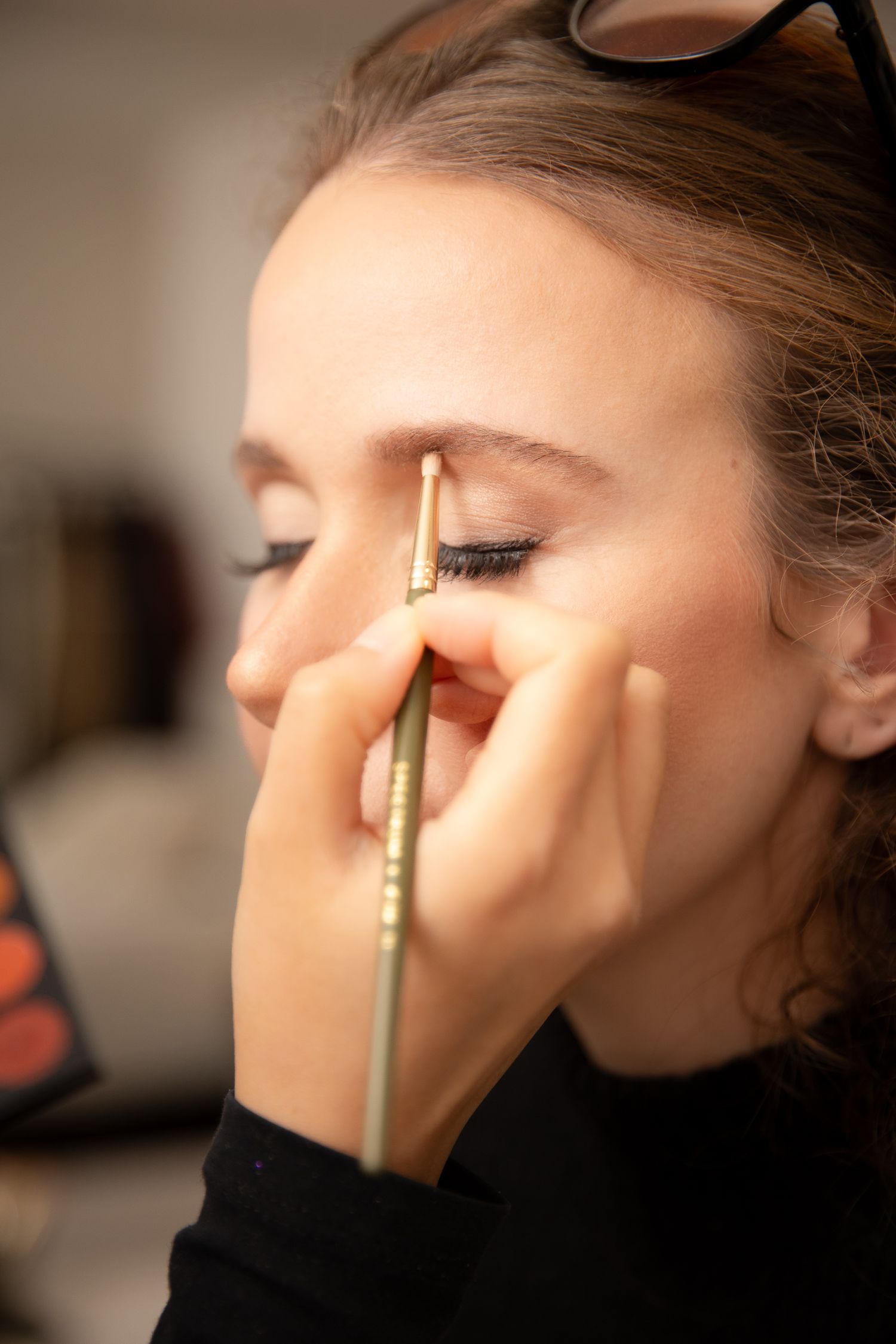 A makeup artist applying eyeliner to a young woman with sunglasses on her head, wearing a black top.